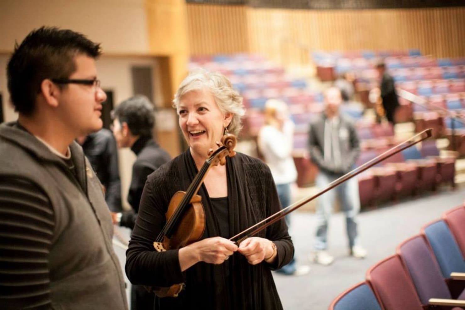 Pam Arnstein in an auditorium, holding a violin and speaking to a student.