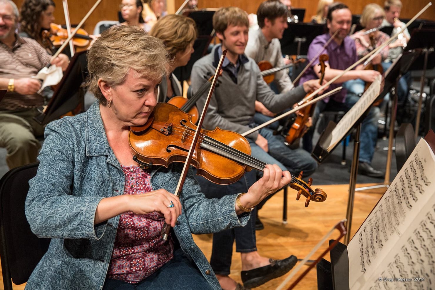 In foreground Pam Arnstein is playing the violin, reading sheet music; behind her are a number of other musicians in conversation or tuning their instruments.