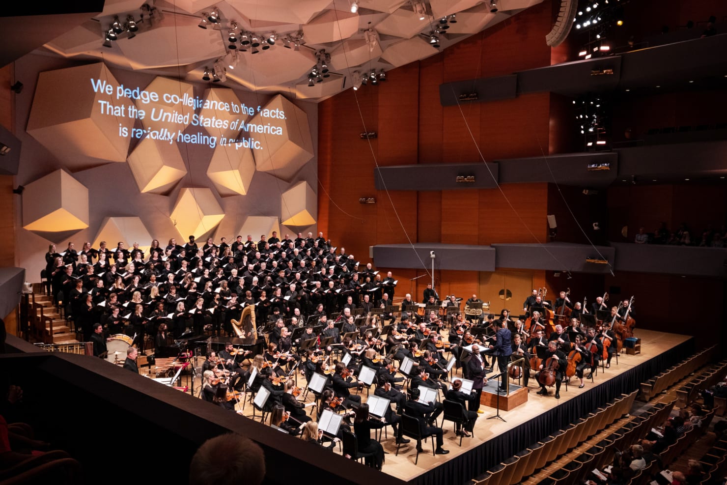 A wide-lens image of the premiere of brea(d)th, showing an 150-person choir and the full Orchestra. The words of the libretto are projected onto the back of the stage.