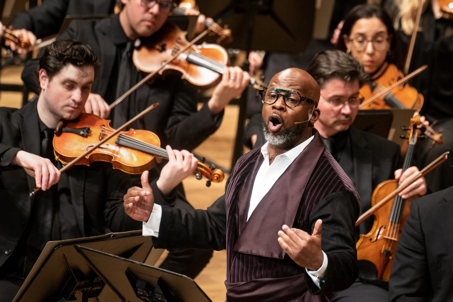 A close-up image of a man performing, gesticulating while a violin section is shown behind him.