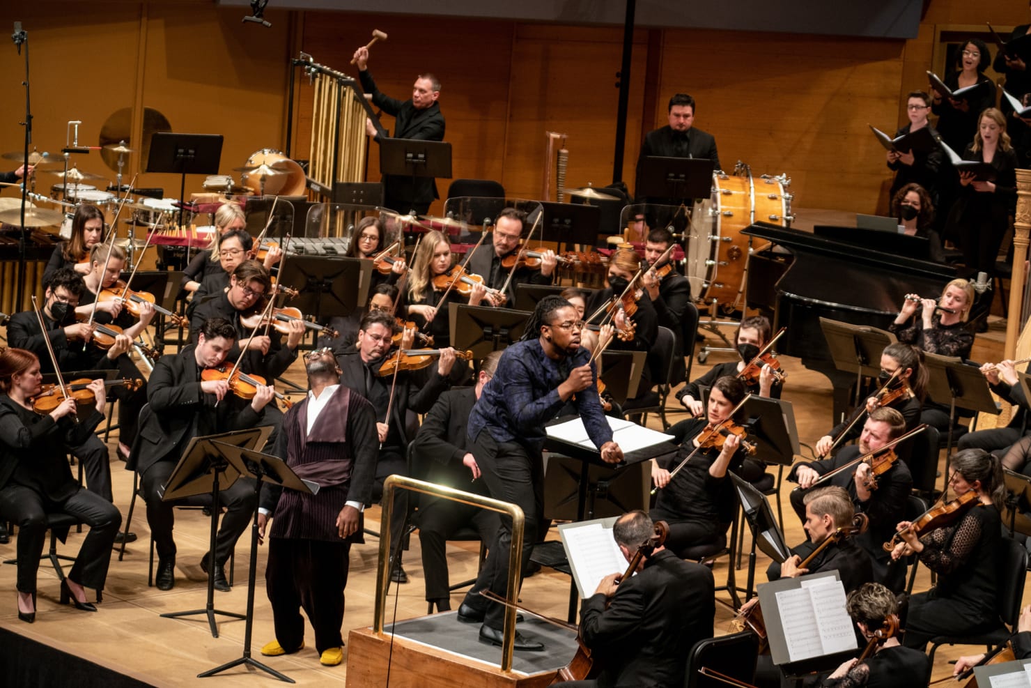 An image of conductor Jonathan Taylor Rush, leaning forward and signaling to the Orchestra; Marc Bamuthi Joseph stands next to the podium.