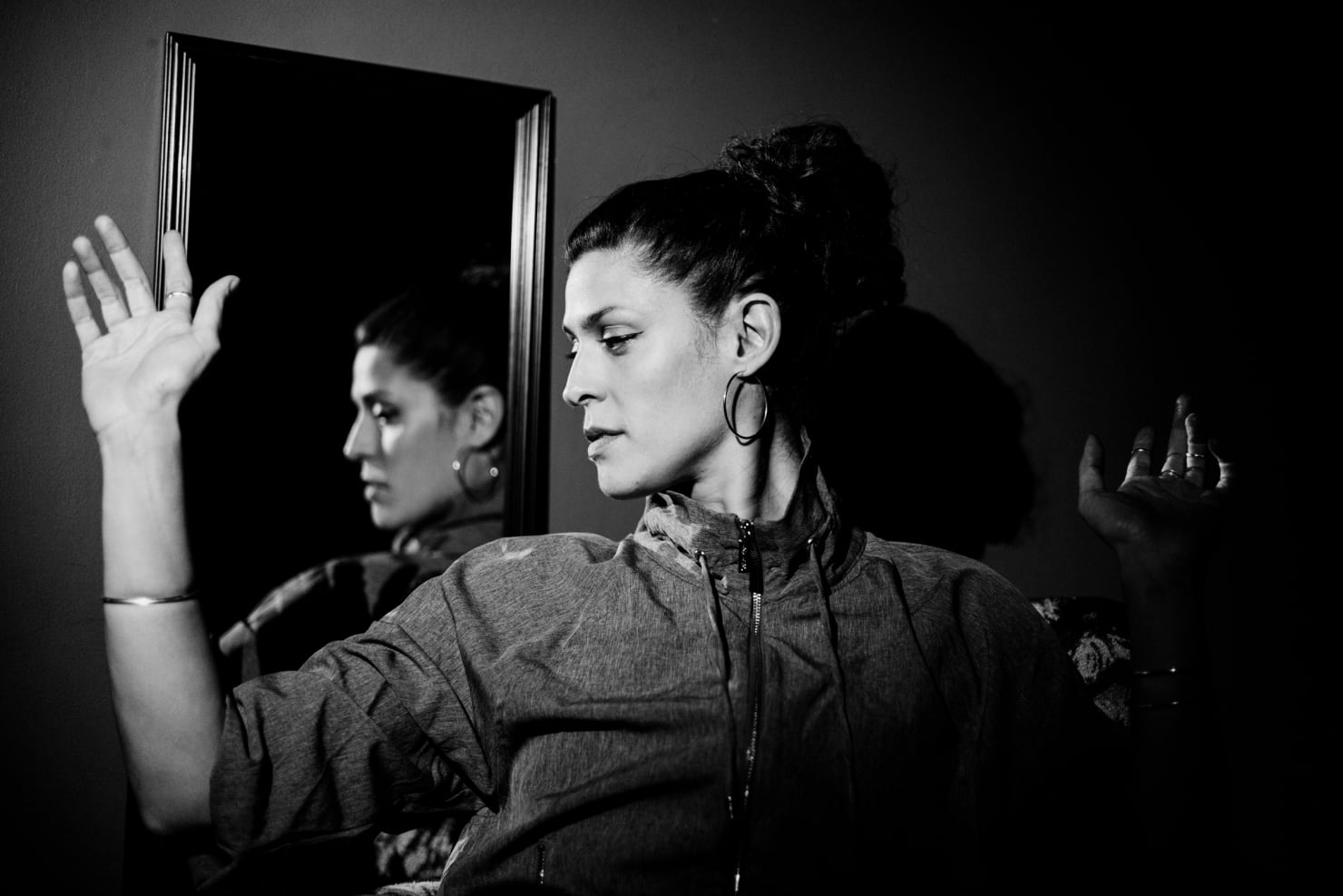 A portrait of Dessa, a woman standing against a wall, holding her arms up. She stands in front of a mirror that her face is reflected in.