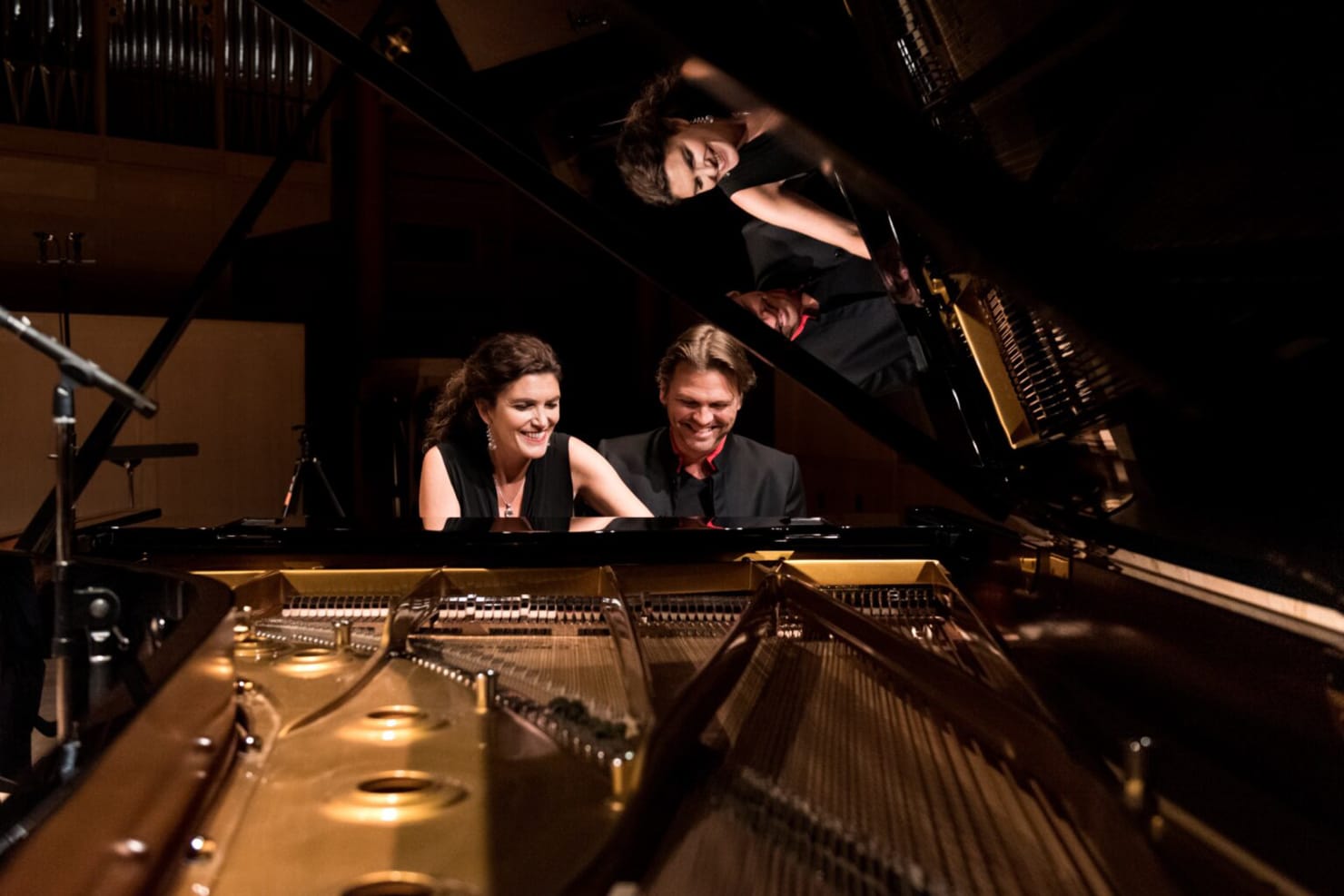 Two people, a woman and a man, sit at piano; the image is from the perspective of the back of the piano, with the whole instrument visible.