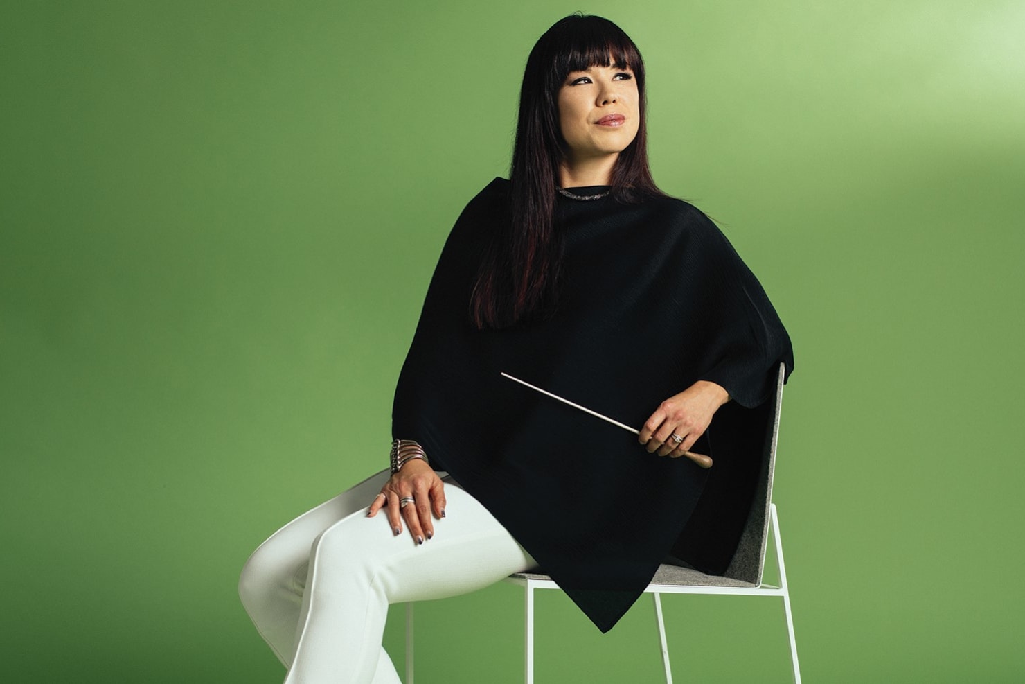 Hicks wearing black and white, sitting on a white chair against a deep green background.