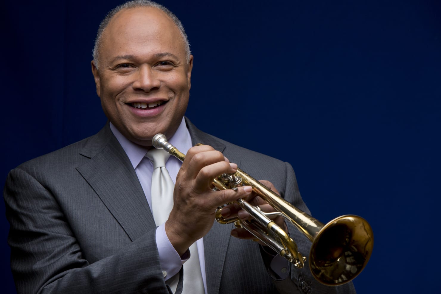 Stripling holding a trumpet and smiling at the camera, against a dark blue background.