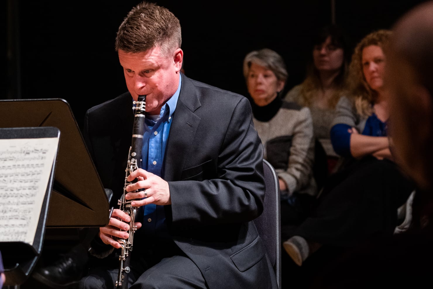 A clarinetist, Greogry T. Williams, blowing into his instrument; a few audience members are positioned behind him.