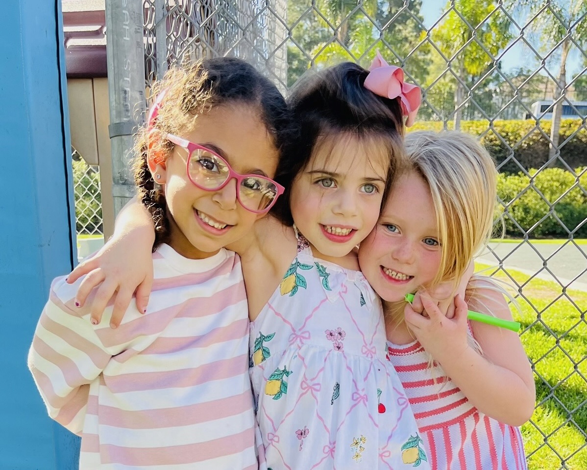 Three girls smiling and standing next to each other, shoulder to shoulder, while standing outside.