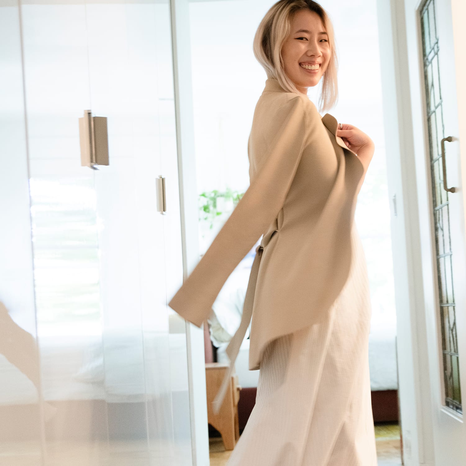 woman wearing tan jacket over her shoulders and long white dress, smiling in an apartment hallway