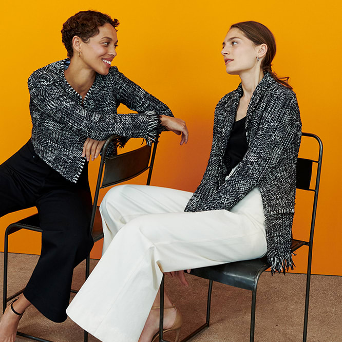 two women sitting in chairs and facing each other, wearing black and white interweave jackets and black and white wide leg jeans