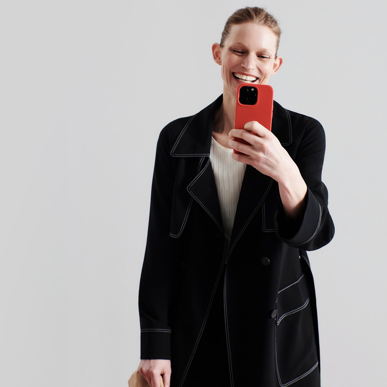 image of woman wearing black trench coat in front of light grey background, and smiling at her phone