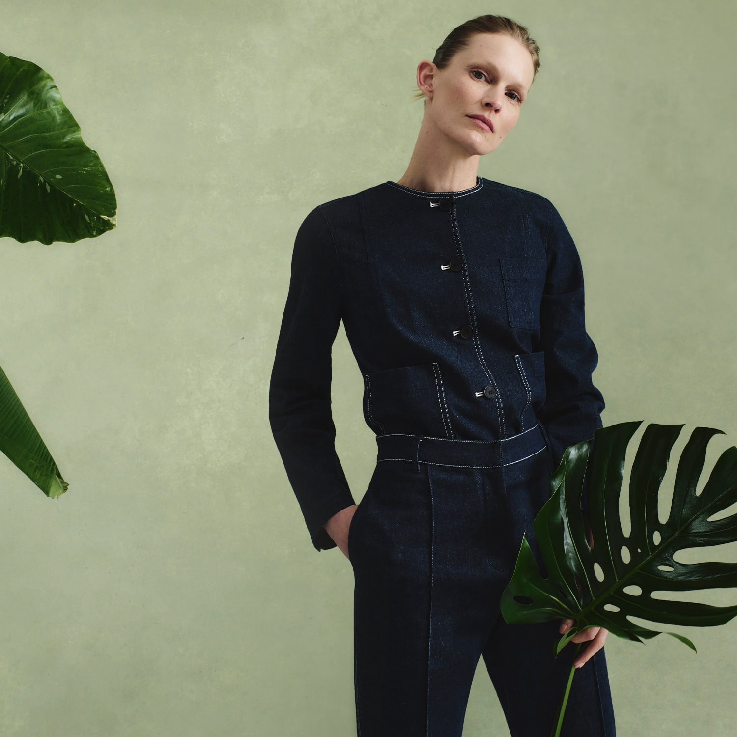 woman wearing dark denim Kane jacket and Christy pant in front of green background with foliage on the sides, holding a monstera leaf