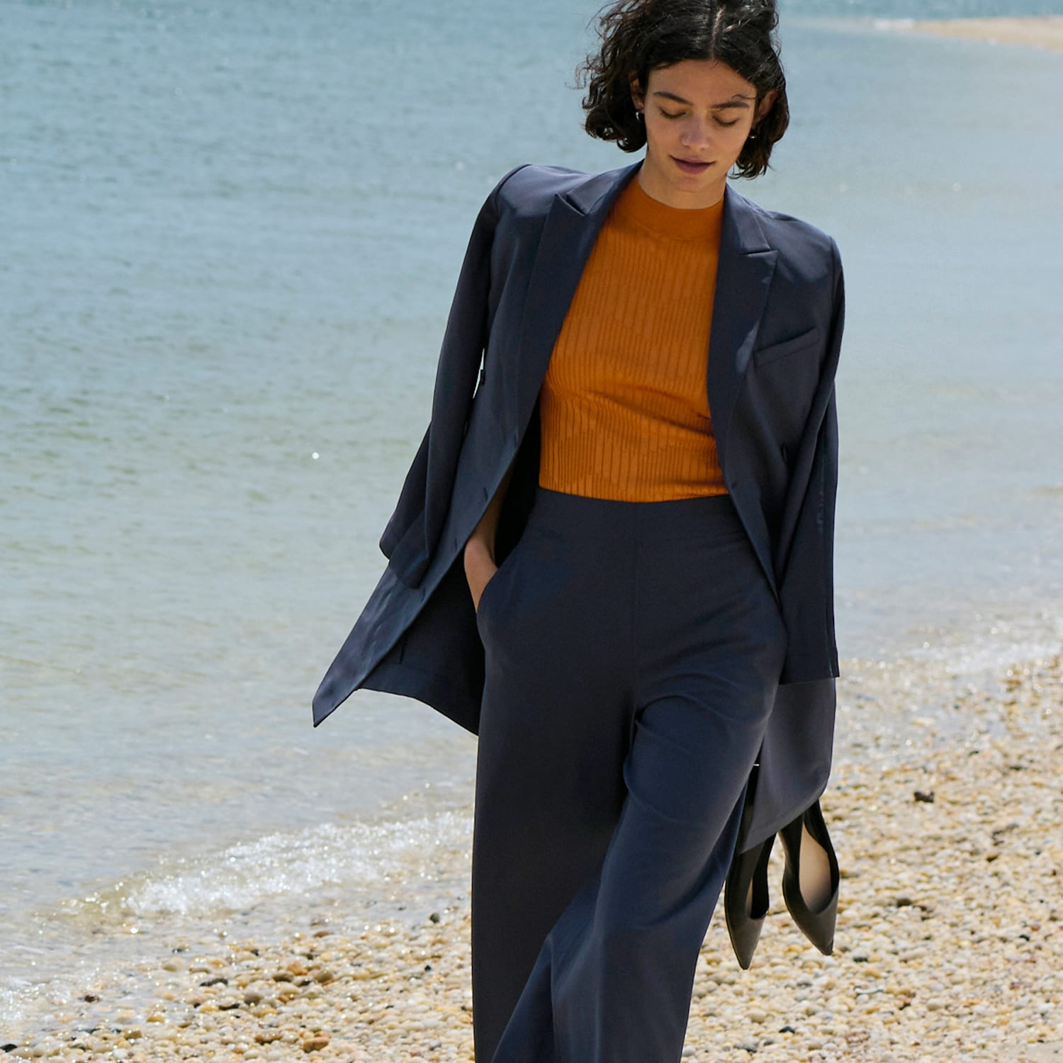 woman wearing cool charcoal colored blazer with orange knit top underneath, holding her shoes in one hand and walking down a beach