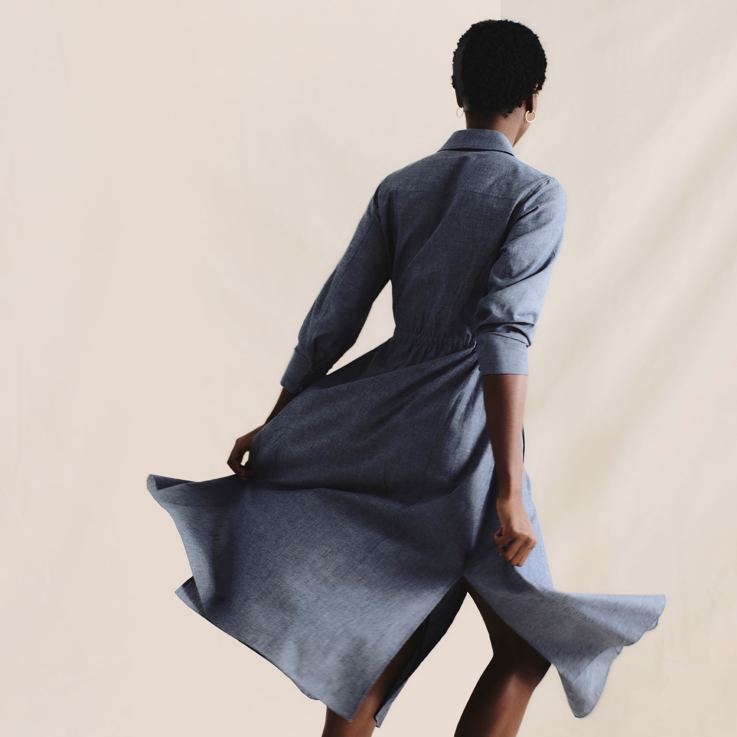 image of a woman facing the back and twirling her blue denim chambray dress in front of a cream-colored curtain backdrop