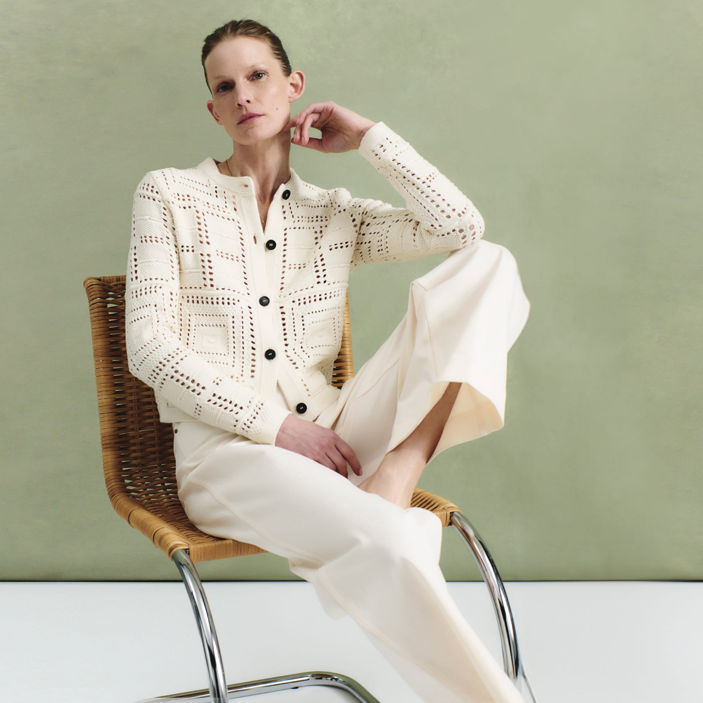 woman wearing ivory crochet-knit cardigan and white jeans, sitting in chair in front of sage green background