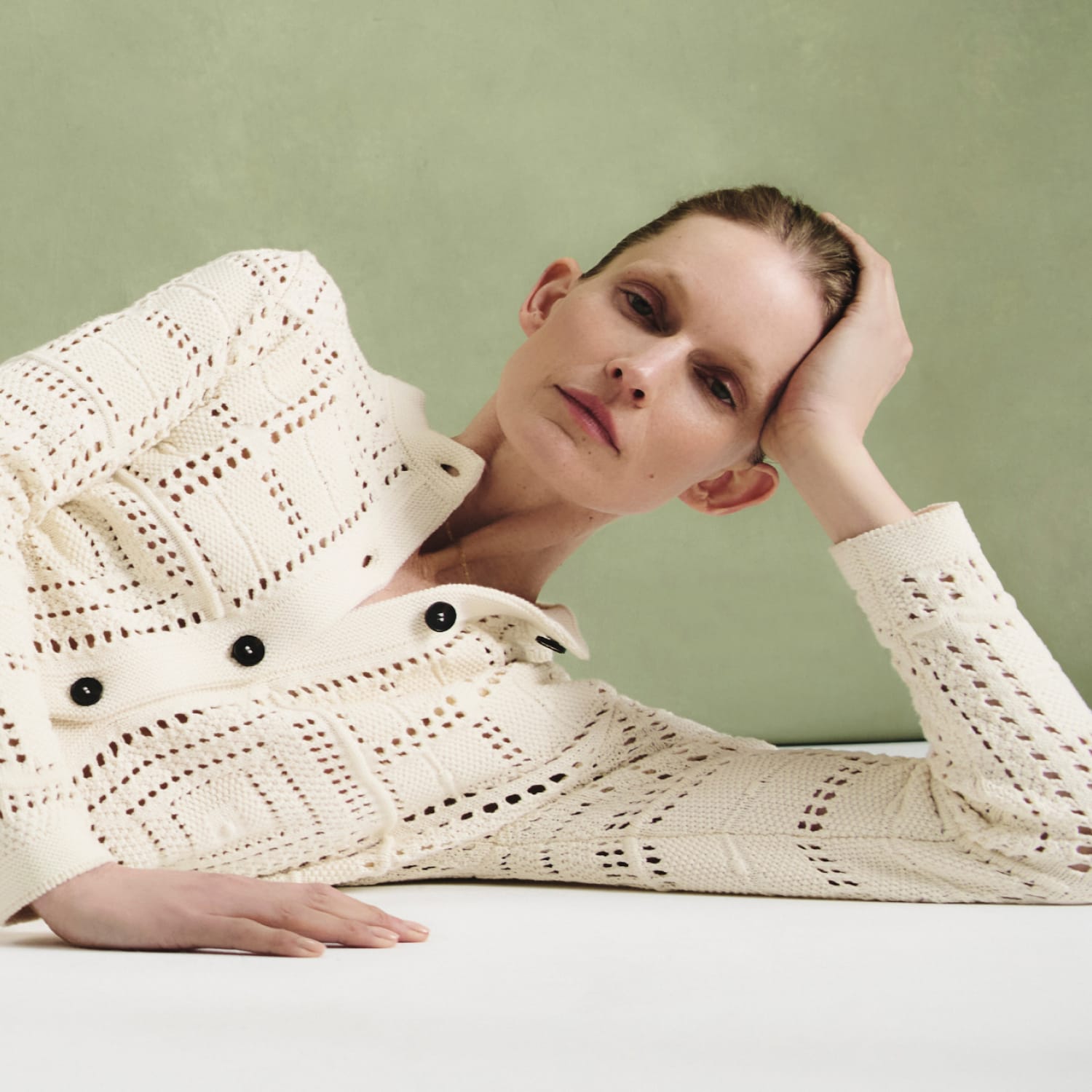 woman lying on white floor with sage green background, wearing ivory crochet-knit cardigan and white jeans