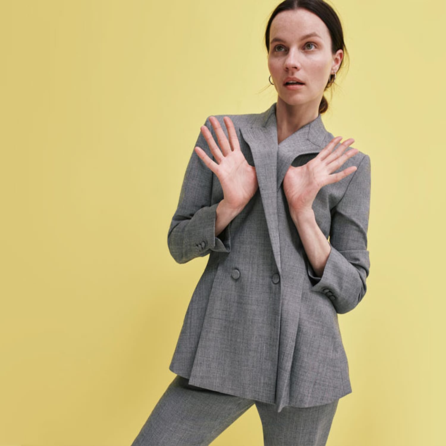 woman lifting collar of Gaia suit blazer in gray sharkskin fabric, standing in front of yellow background