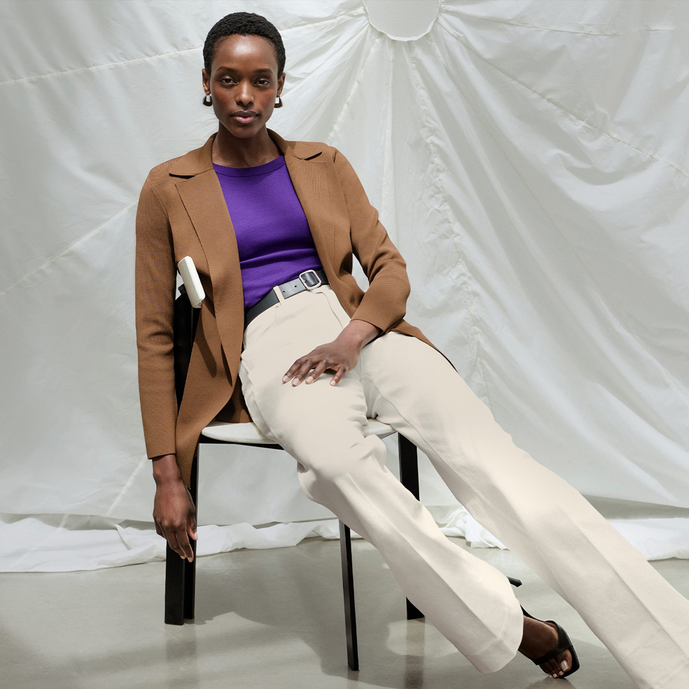 woman wearing Merritt jardigan in saddle color, Milo wide-leg jeans in bright ivory color, sitting in a chair in front of white parachute background and extending her legs out