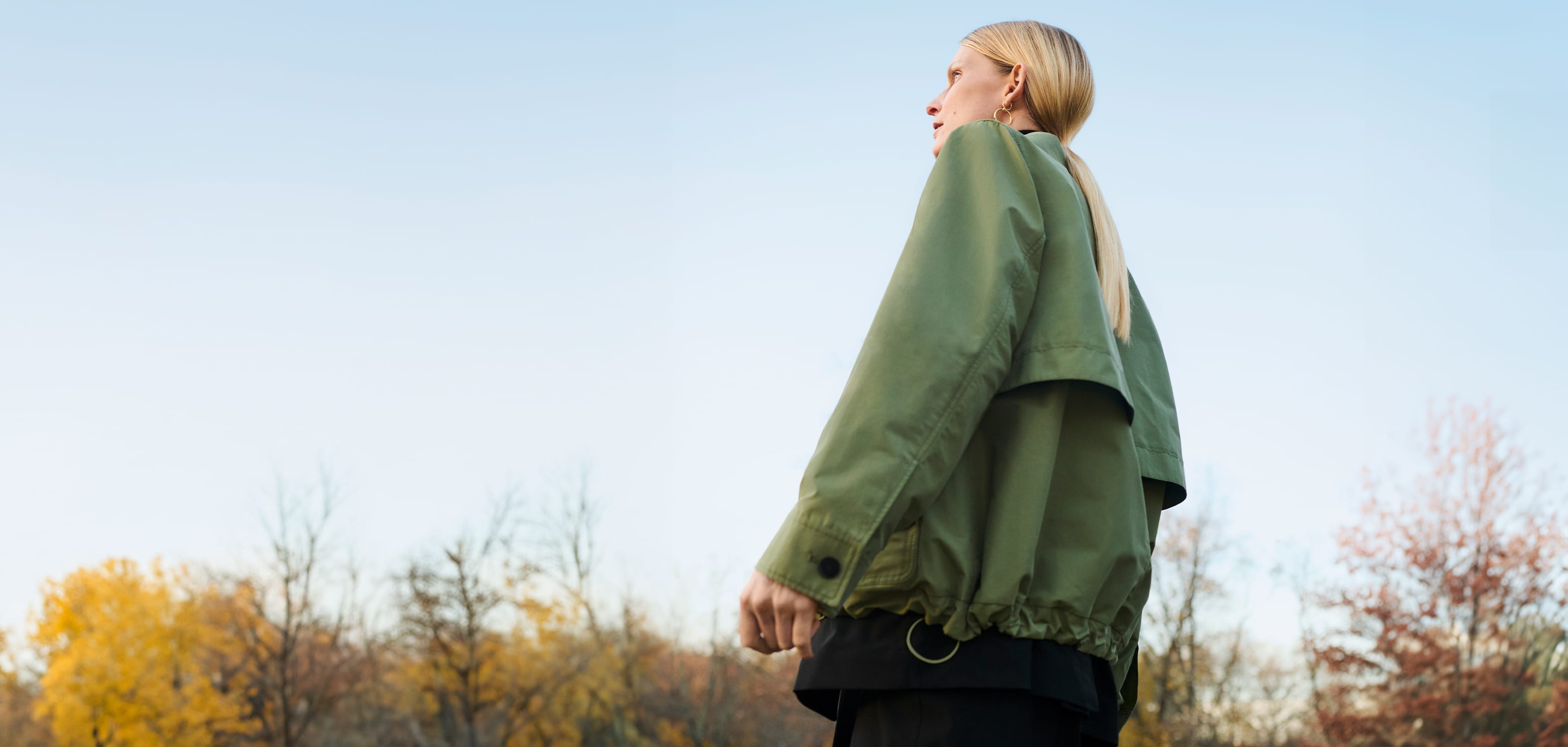 woman with blond ponytail with her back to camera looking into distance, wearing green outerwear jacket