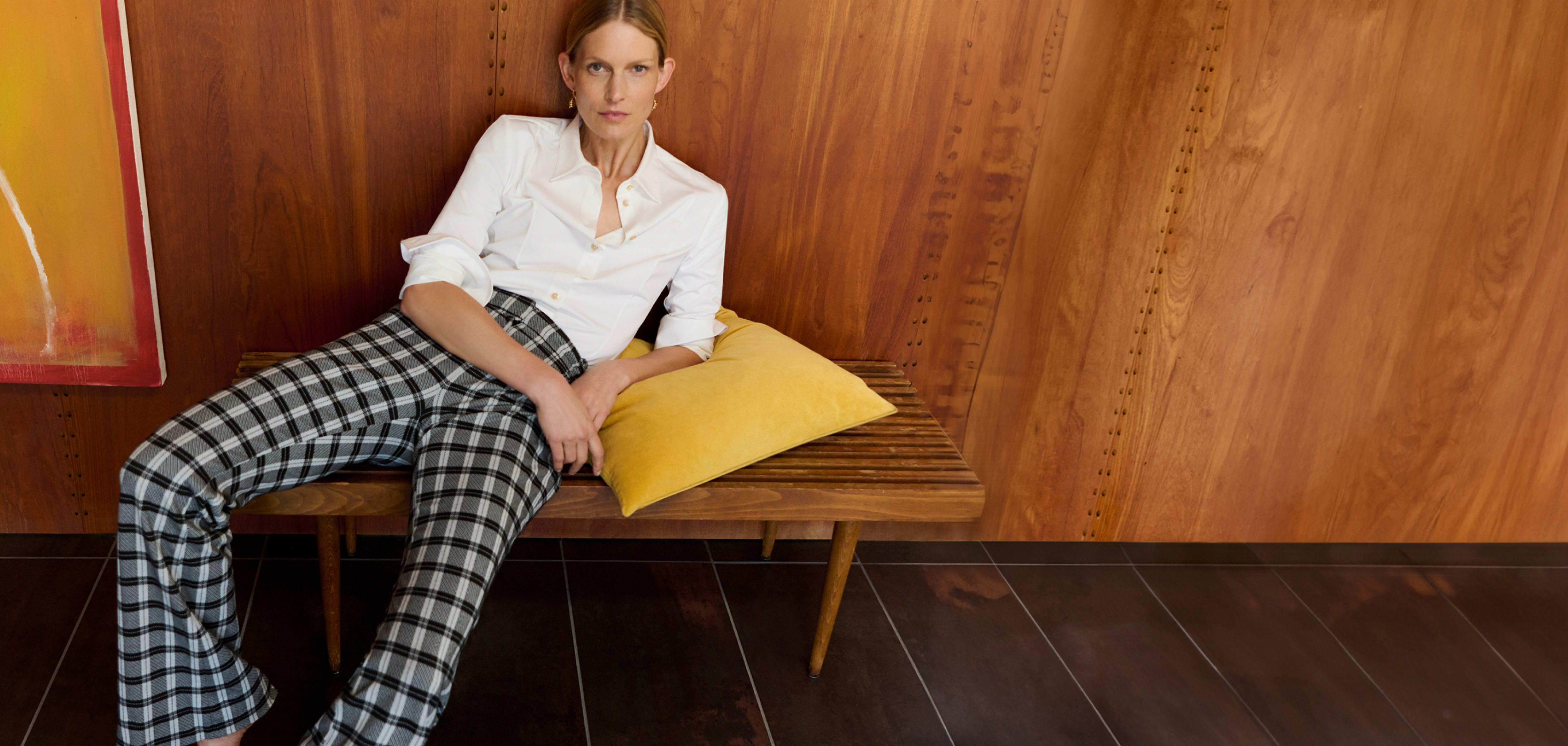 woman lounging on bench with yellow pillow, wearing white button-down top and black/white checkered pants