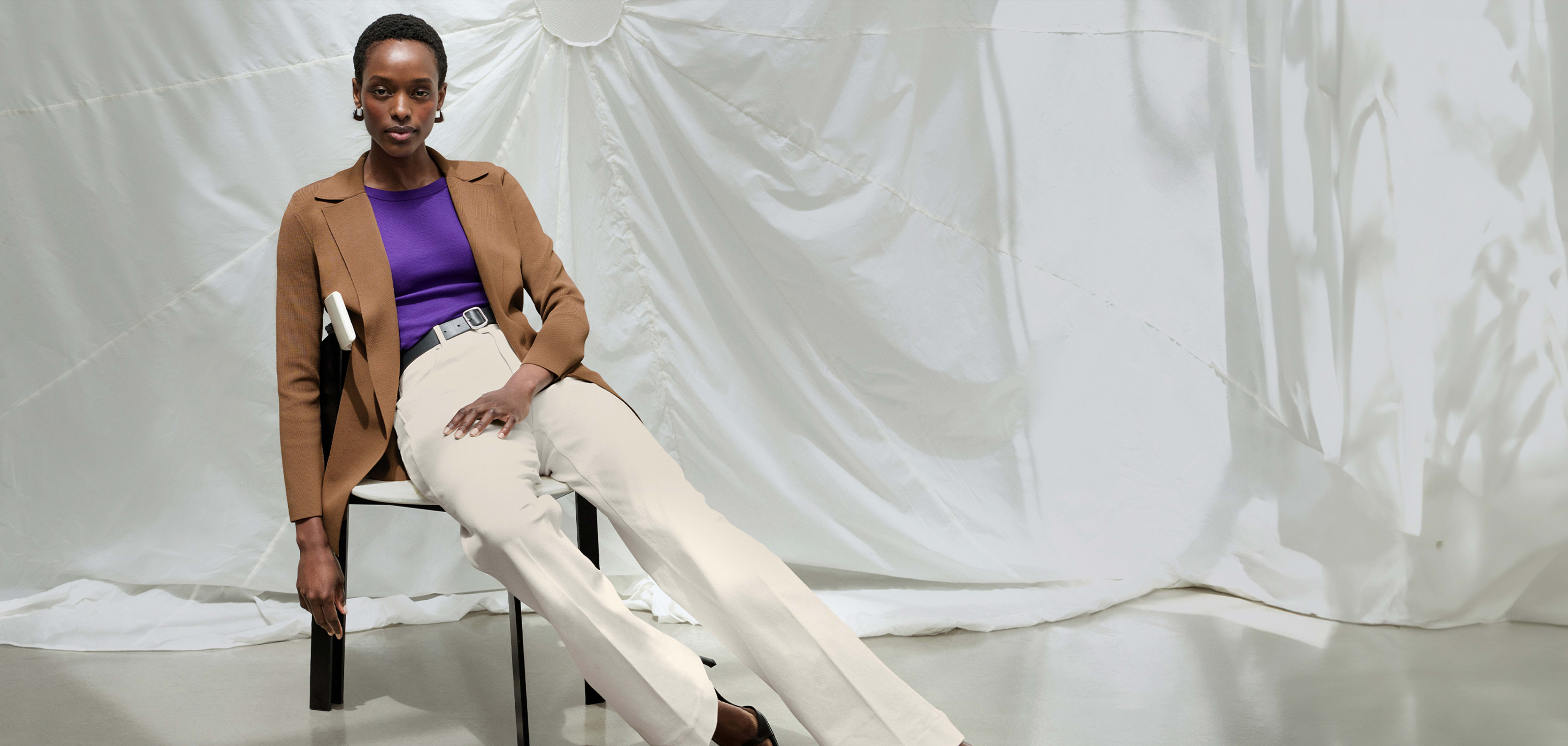 woman wearing Merritt jardigan in saddle color, Milo wide-leg jeans in bright ivory color, sitting in a chair in front of white parachute background and extending her legs out