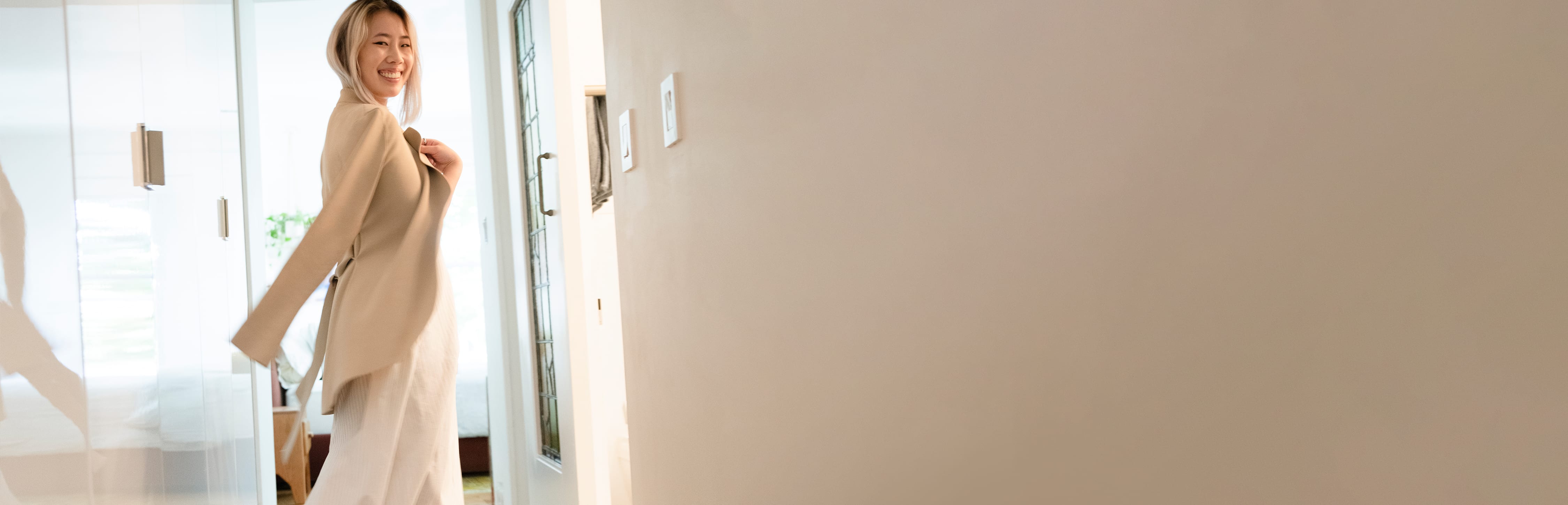 woman wearing tan jacket over her shoulders and long white dress, smiling in an apartment hallway