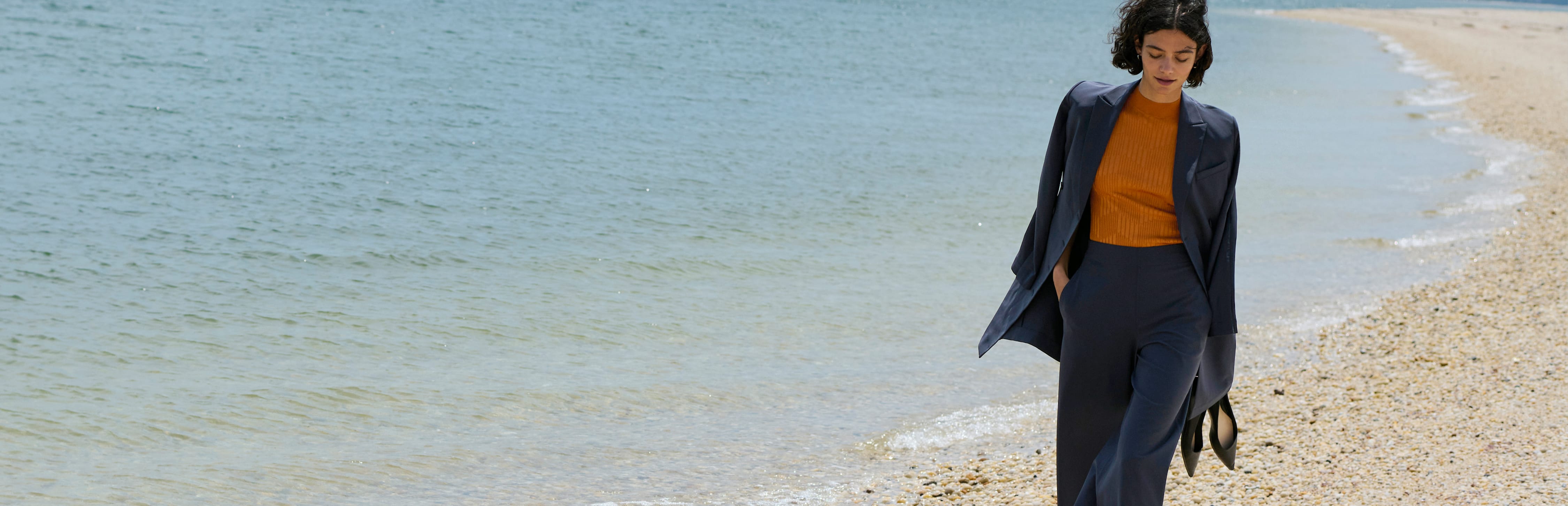 woman wearing cool charcoal colored blazer with orange knit top underneath, holding her shoes in one hand and walking down a beach