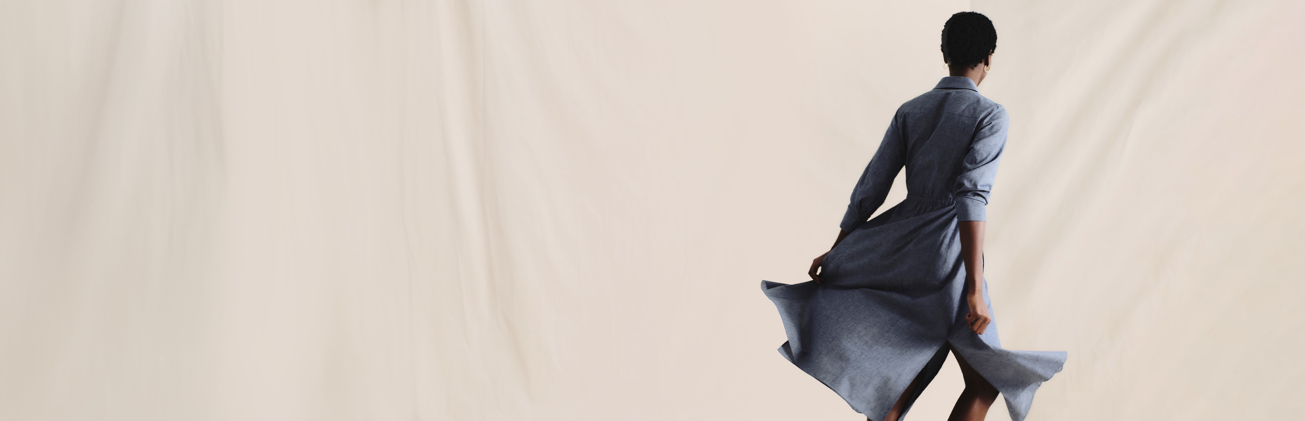 image of a woman facing the back and twirling her blue denim chambray dress in front of a cream-colored curtain backdrop