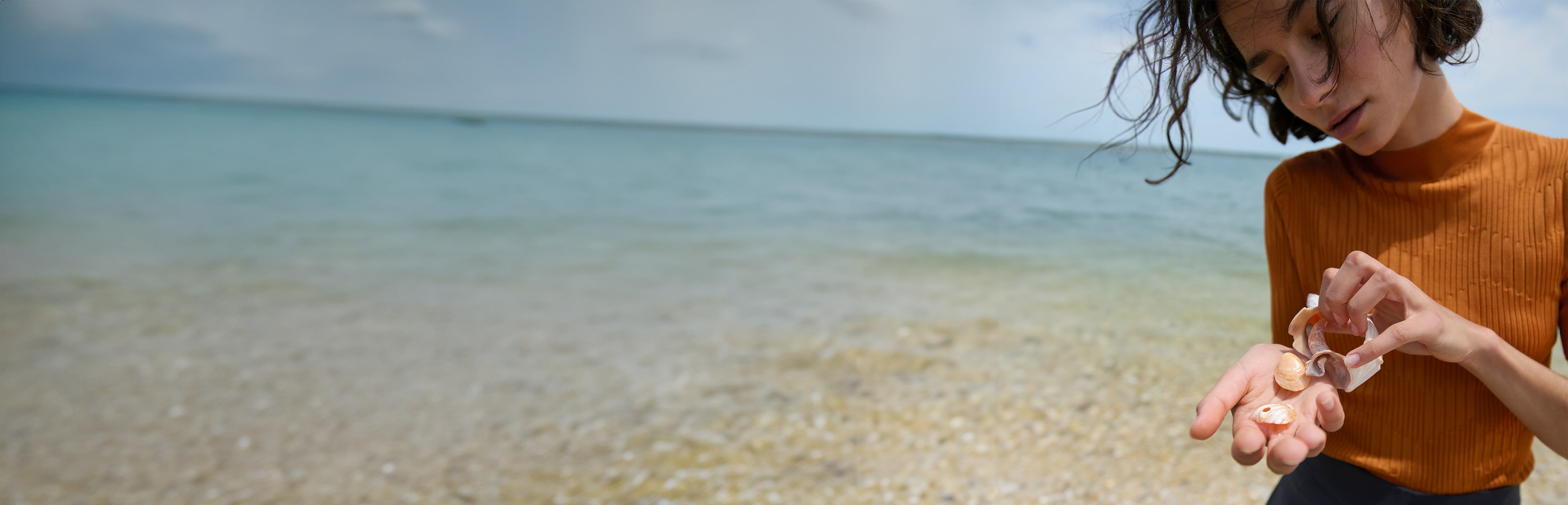 woman looking at seashells in her hand on the beach, wearing orange short sleeve knit Montean top