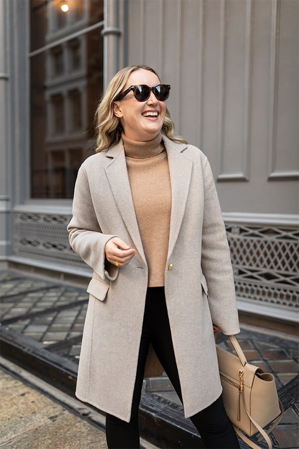 woman standing wearing a beige coat over a camel turtle neck and black pants