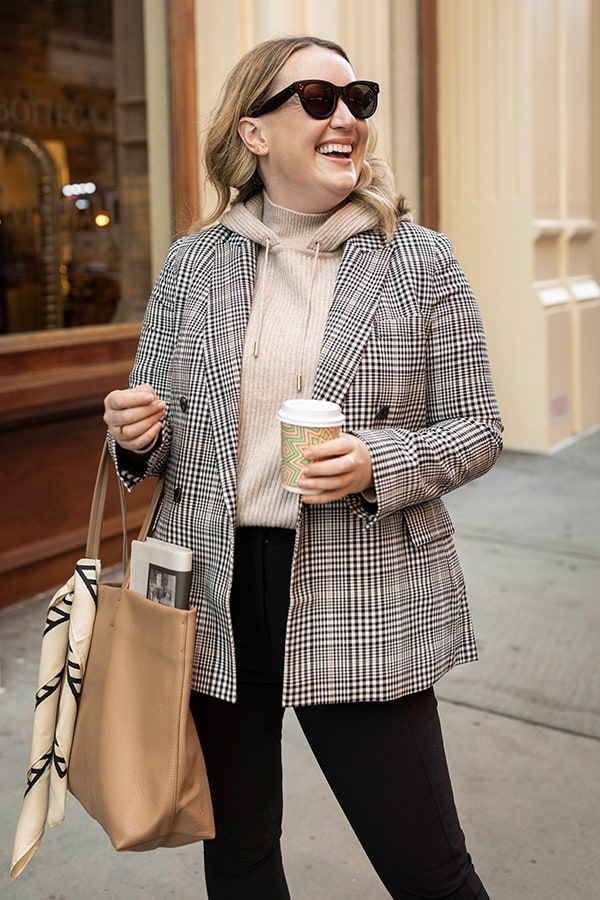 woman standing with a coffee and a tote wearing a hoodie and blazer
