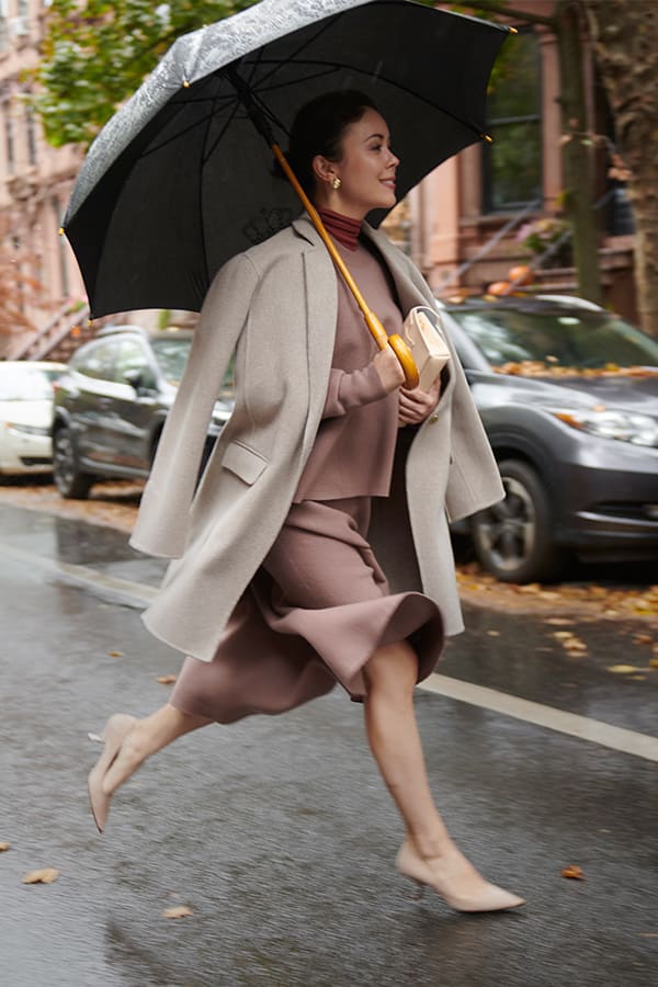 image of a woman wearing a rosy taupe skirt and top under a light gray coat running through the rain with an umbrella