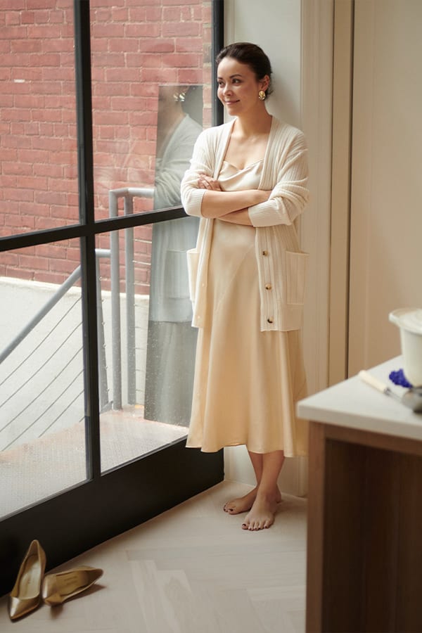 image of a woman looking out her window wearing a cream cardigan over a champagne colored dress