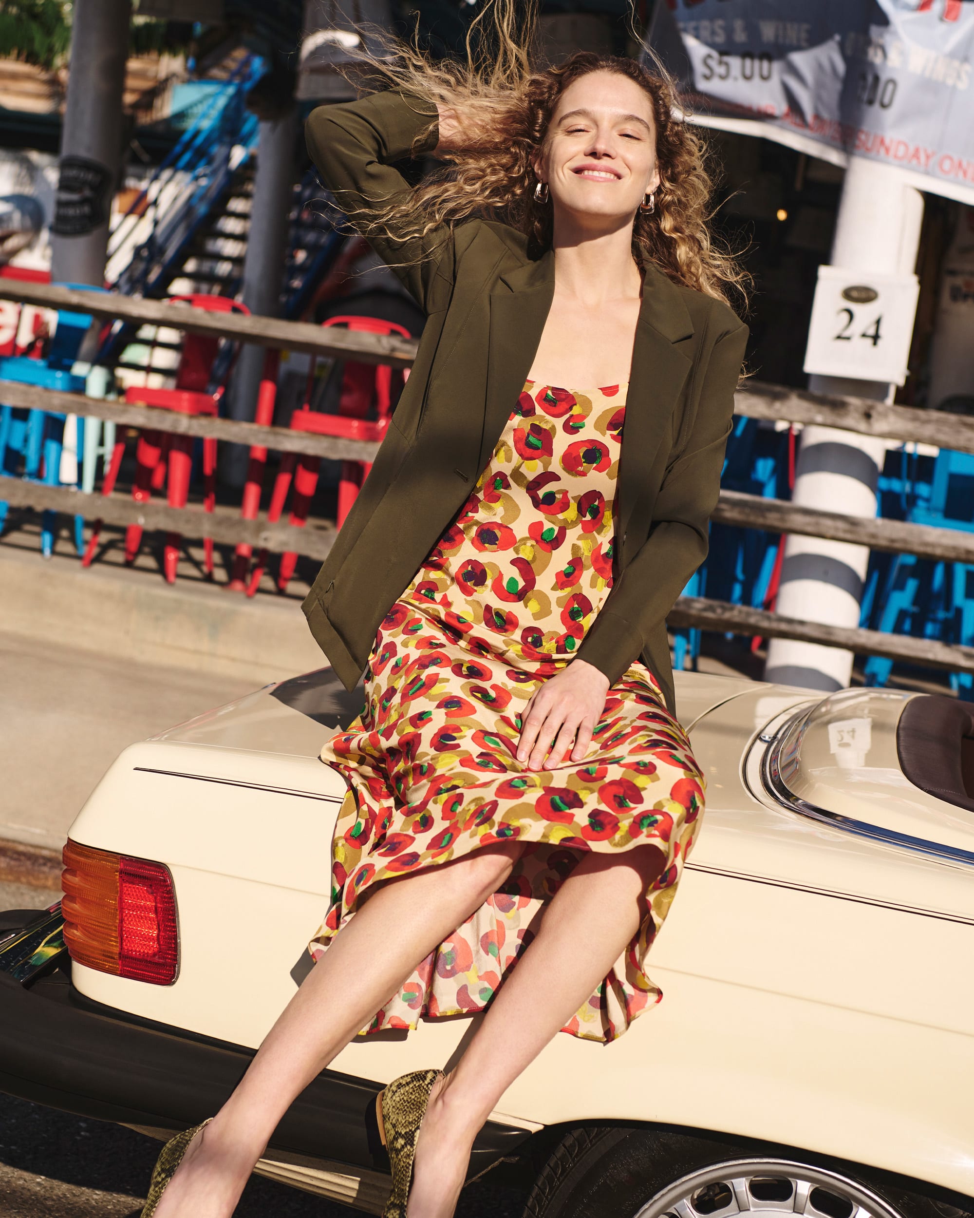 image of a woman sitting on a car wearing the nene dress in peacock and moreland jacket in olive