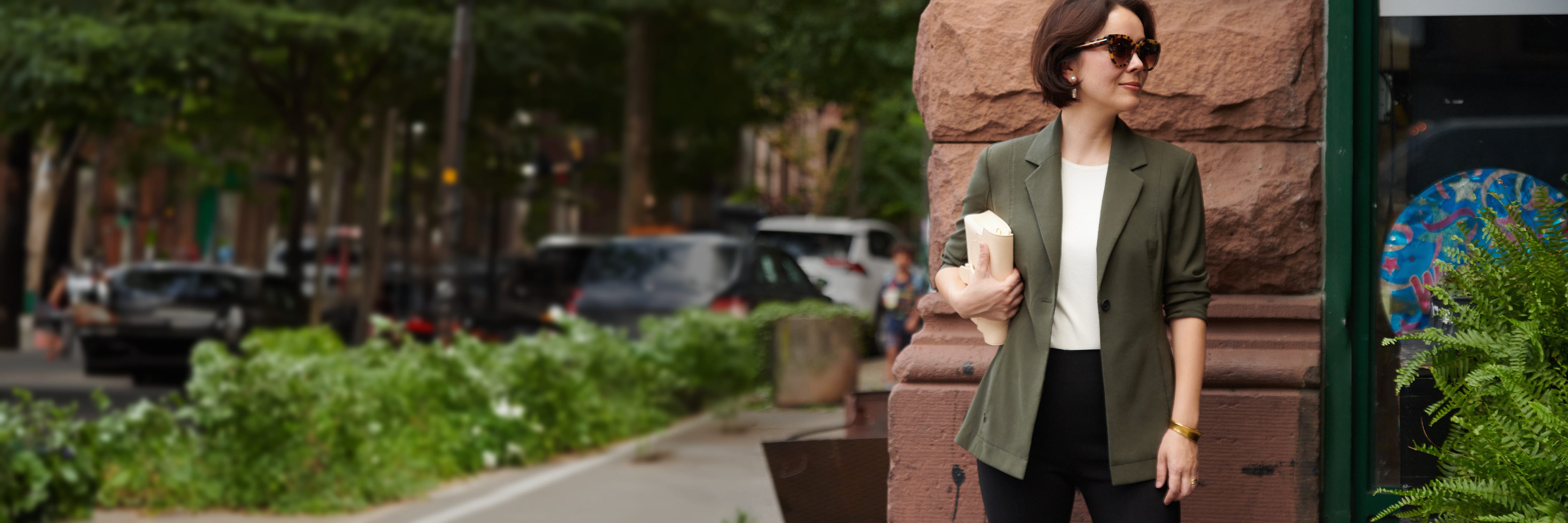 Sarah LaFleur, wearing the Moreland jacket in olive and sunglasses, carrying a cream-colored handbag and standing on a street corner in Brooklyn