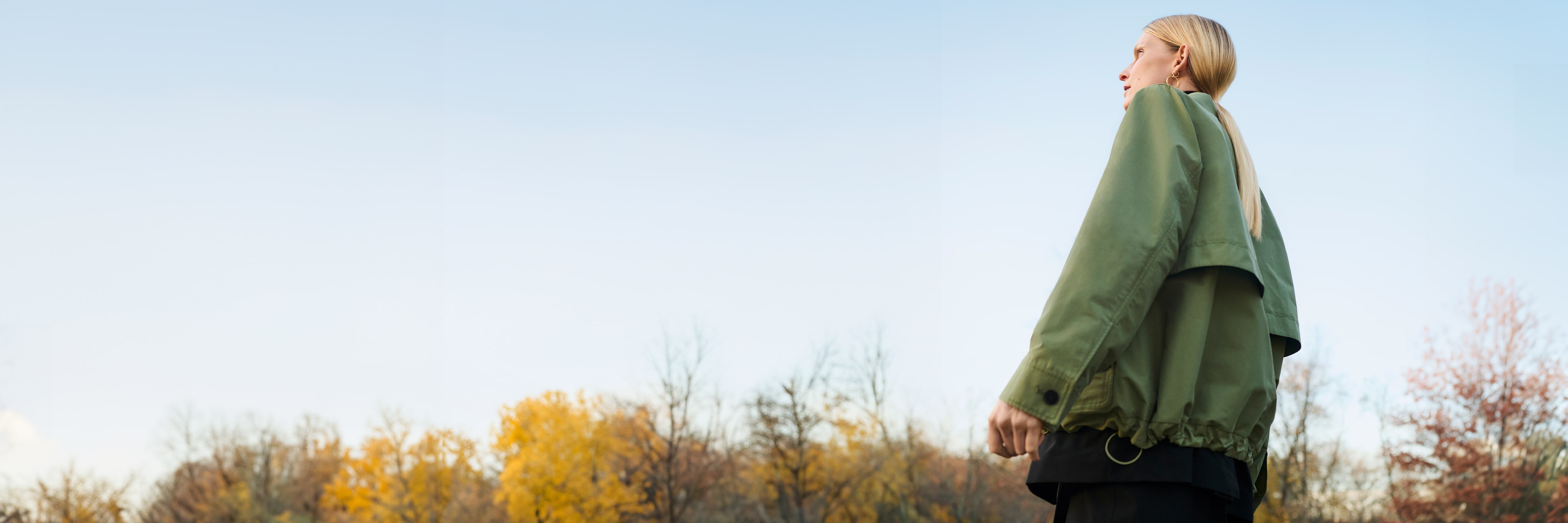 woman with blond ponytail with her back to camera looking into distance, wearing green outerwear jacket