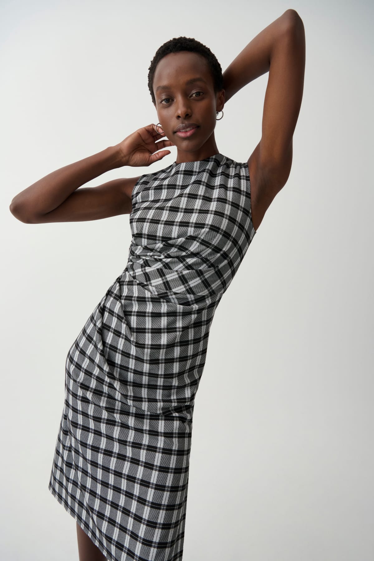 woman in front of light grey background with arms above her head, wearing black/white check plaid sleeveless dress