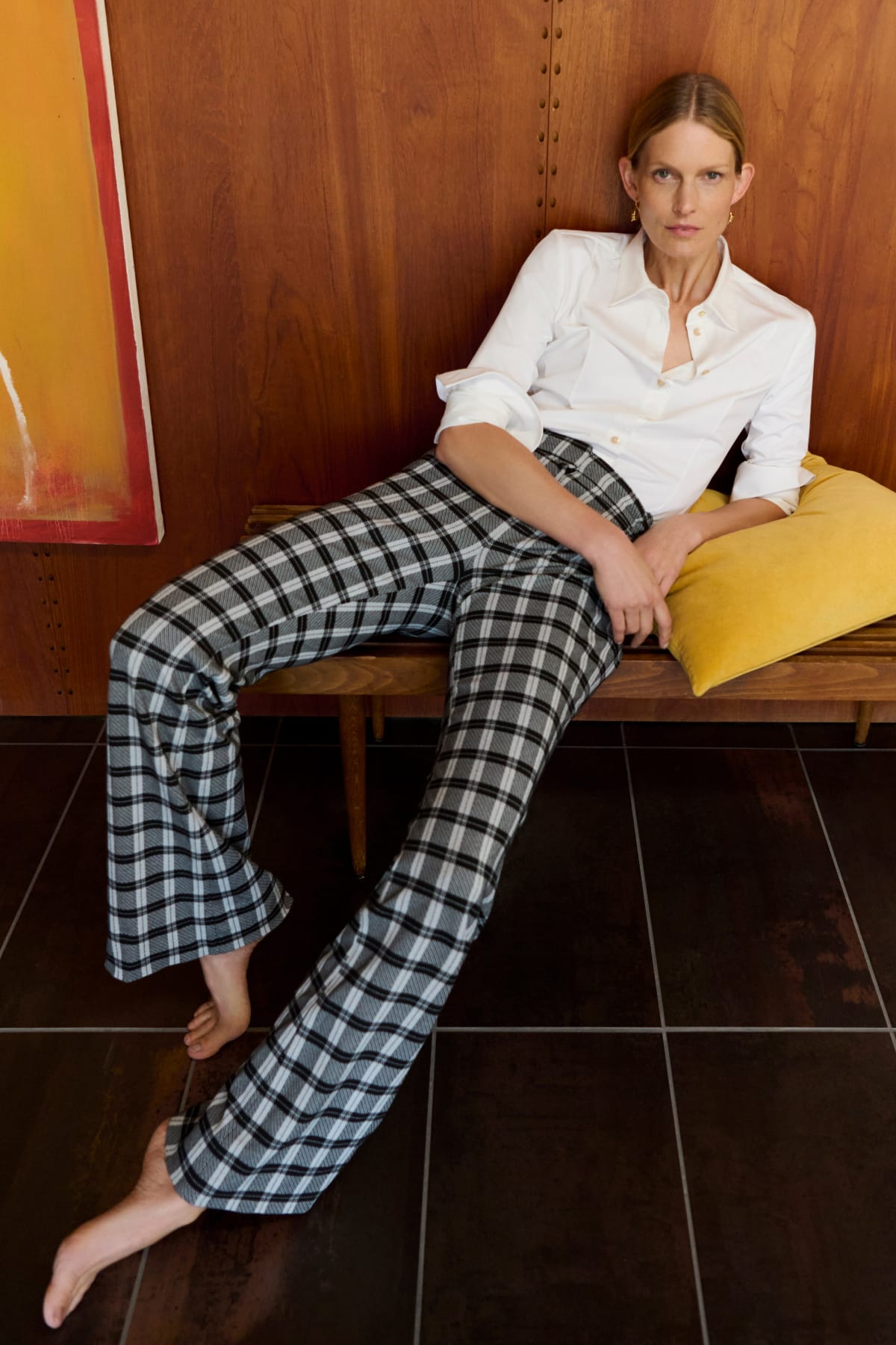 woman lounging on bench with yellow pillow, wearing white button-down top and black/white checkered pants