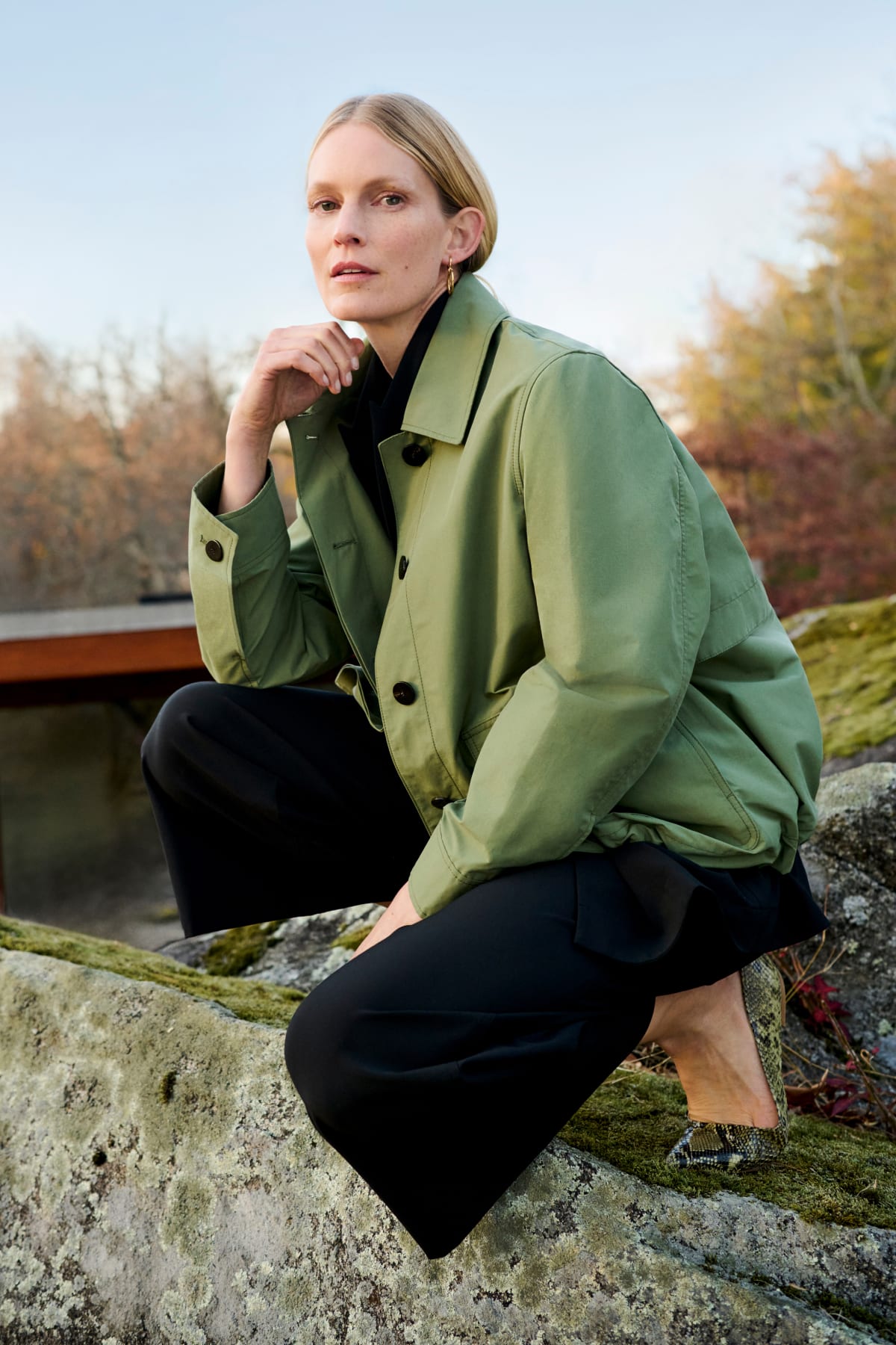 woman crouching on rocks outdoors, wearing light green outerwear jacket and black pants