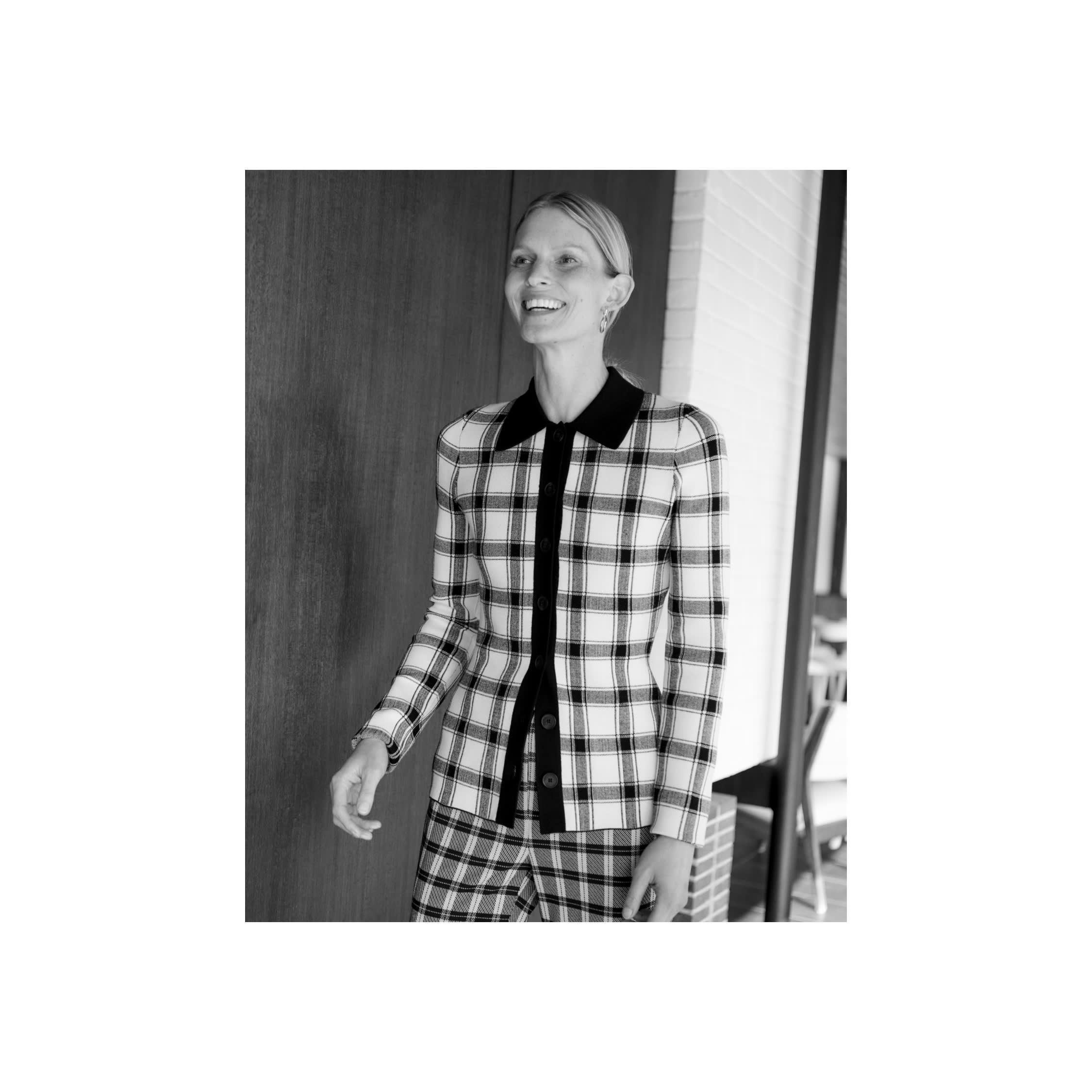 black/white image of woman standing indoors and smiling and wearing black/white knit plaid button-up top