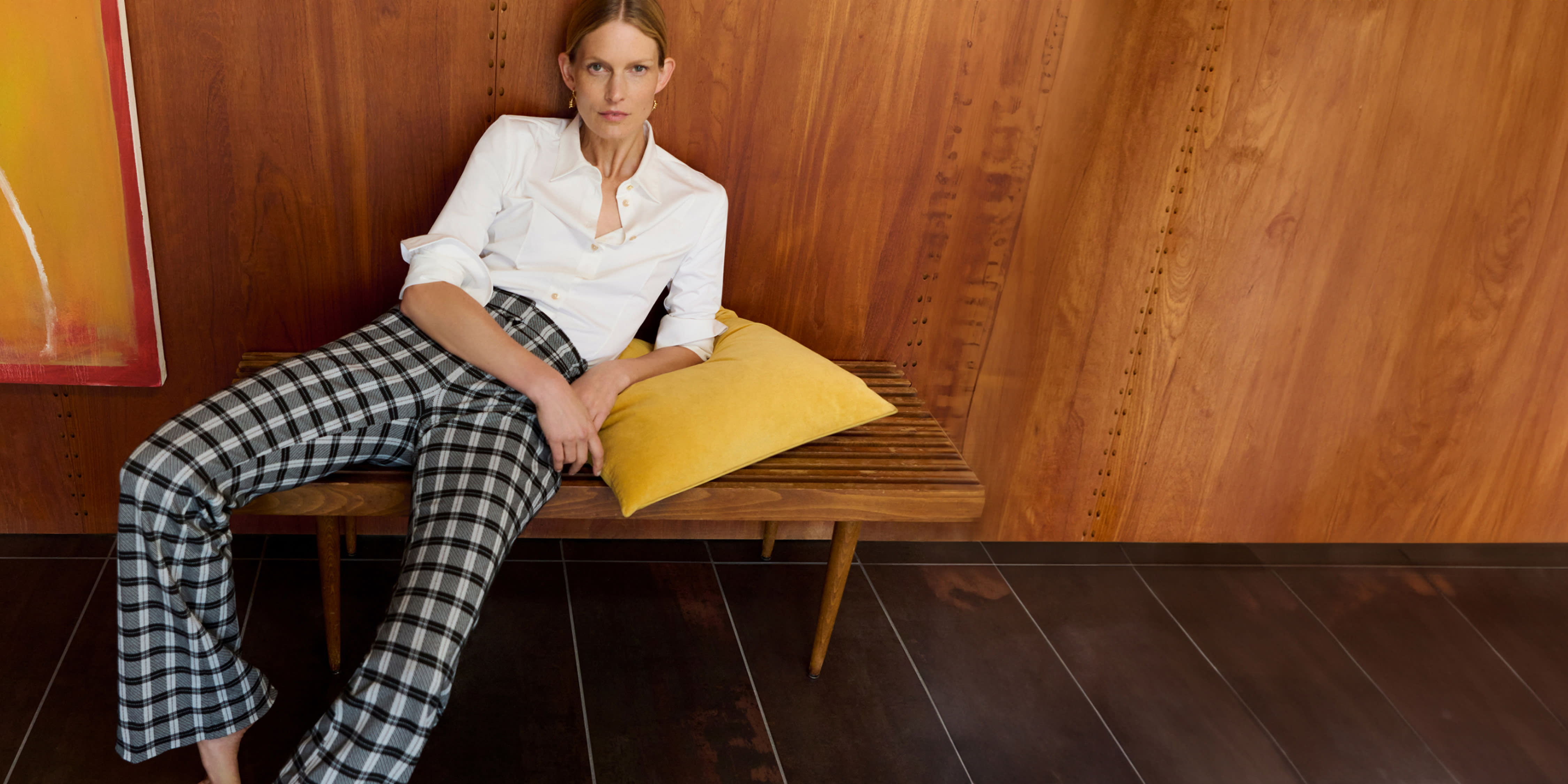 woman lounging on bench with yellow pillow, wearing white button-down top and black/white checkered pants