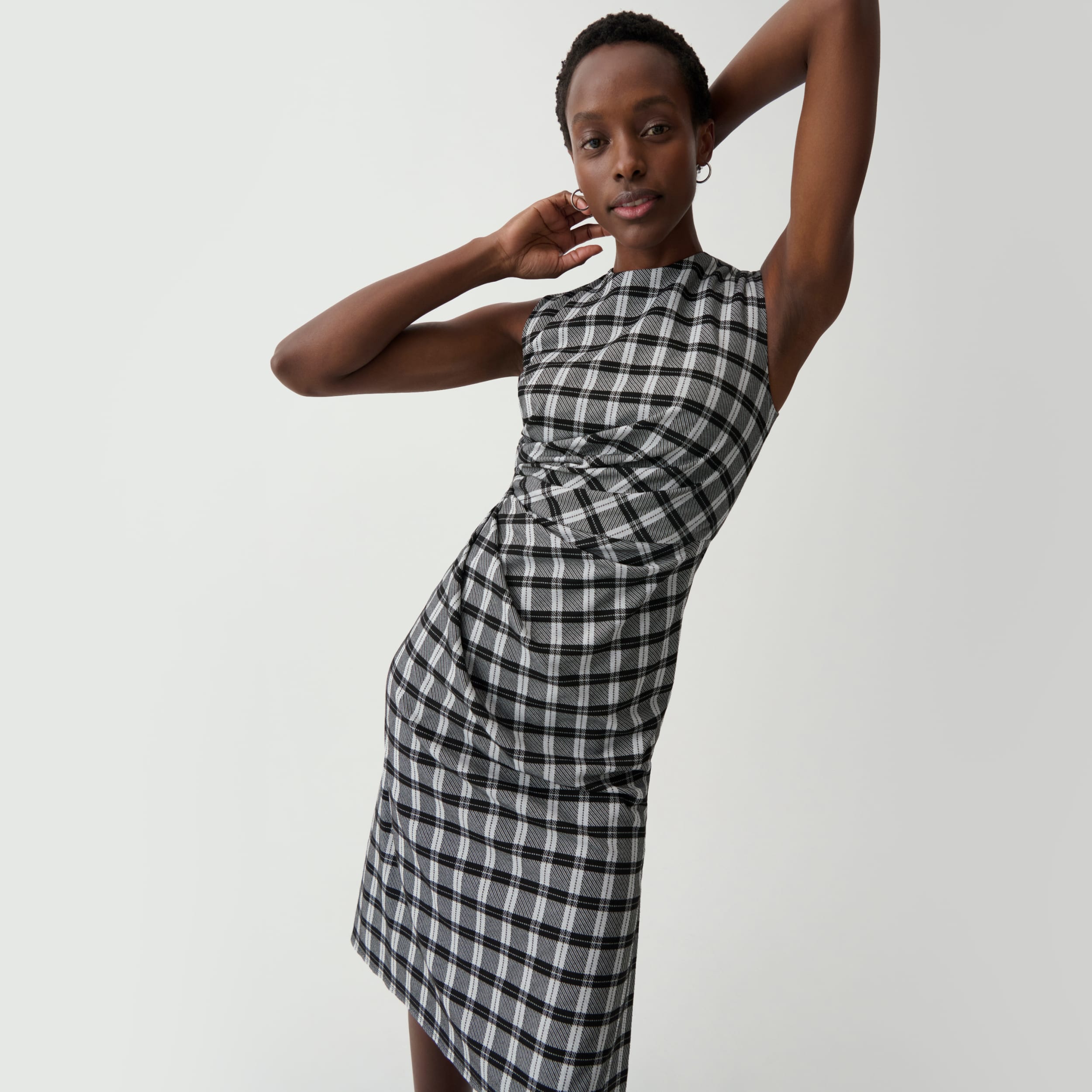 woman in front of light grey background with arms above her head, wearing black/white check plaid sleeveless dress