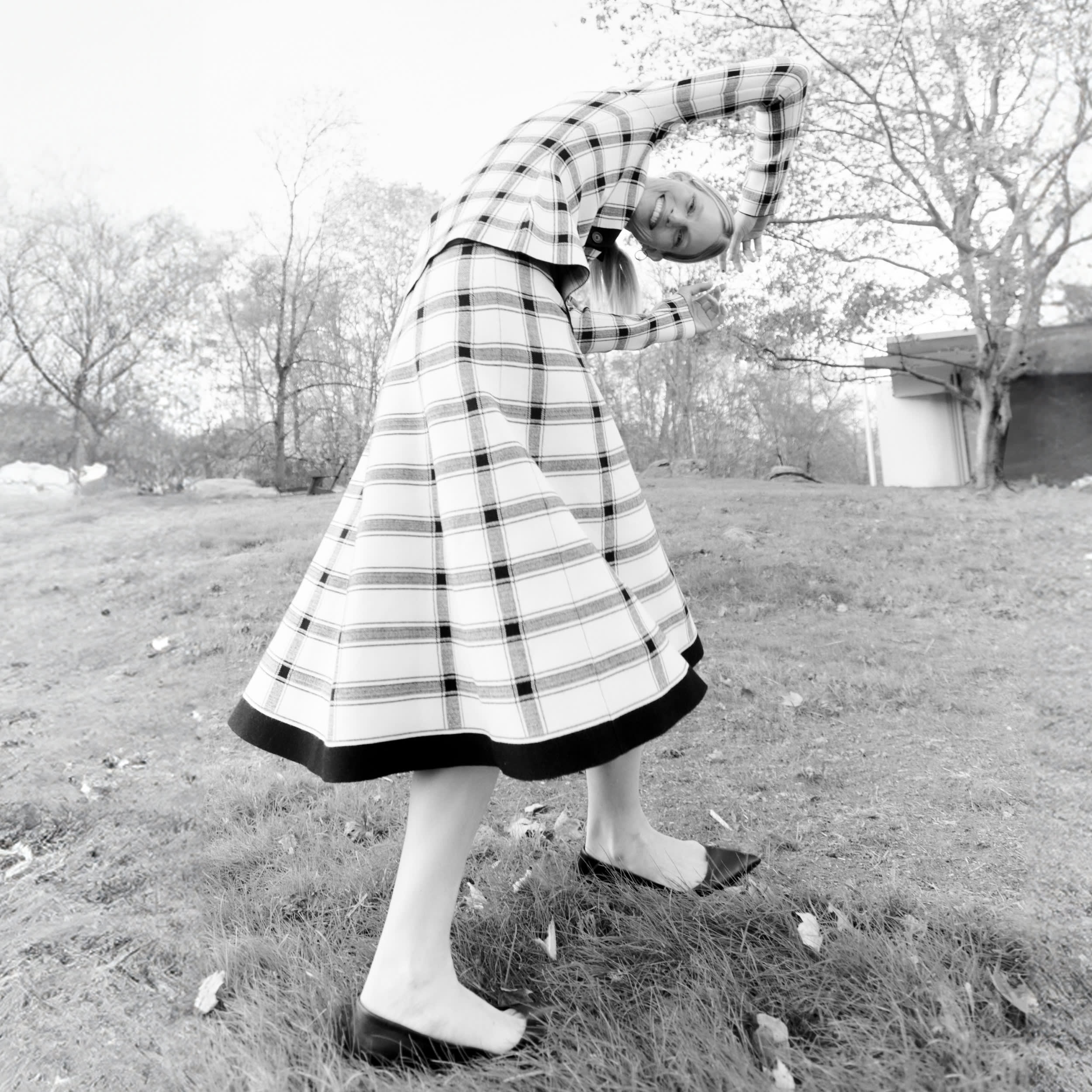 black/white image of woman wearing black/white knit plaid top and skirt set, bending over with her arms above her head outside