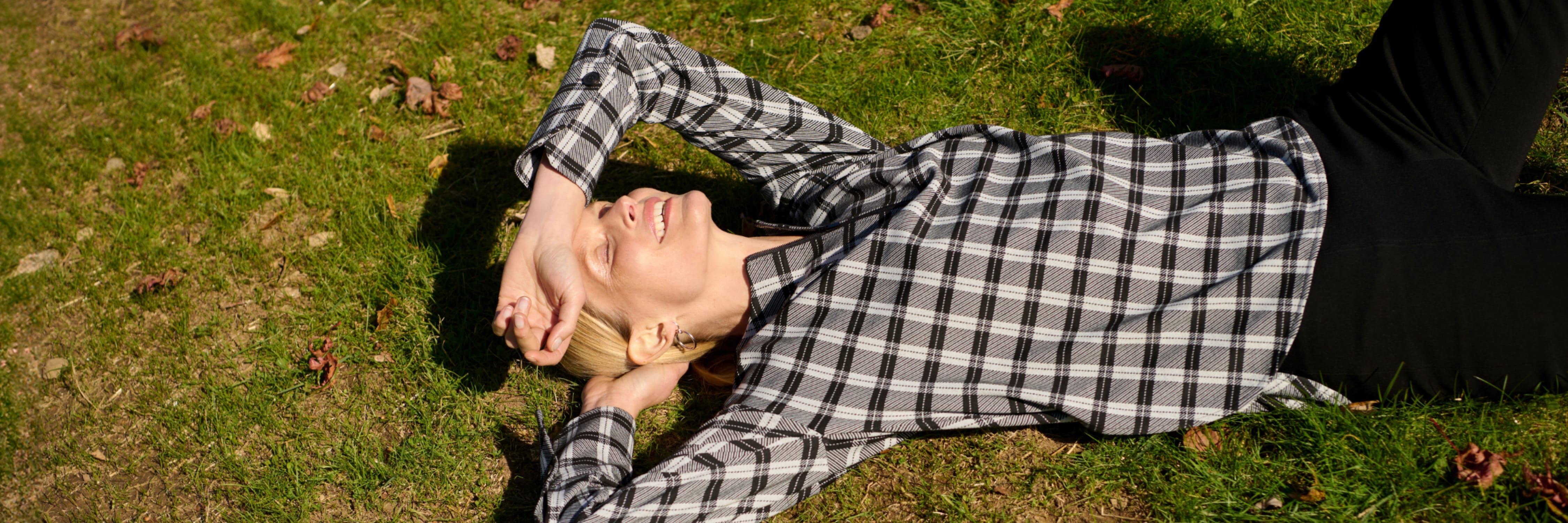 smiling woman lying down in grass with one hand supporting her head and the other arm raised up to cover her forehead; wearing black/white check pattern top and black pants