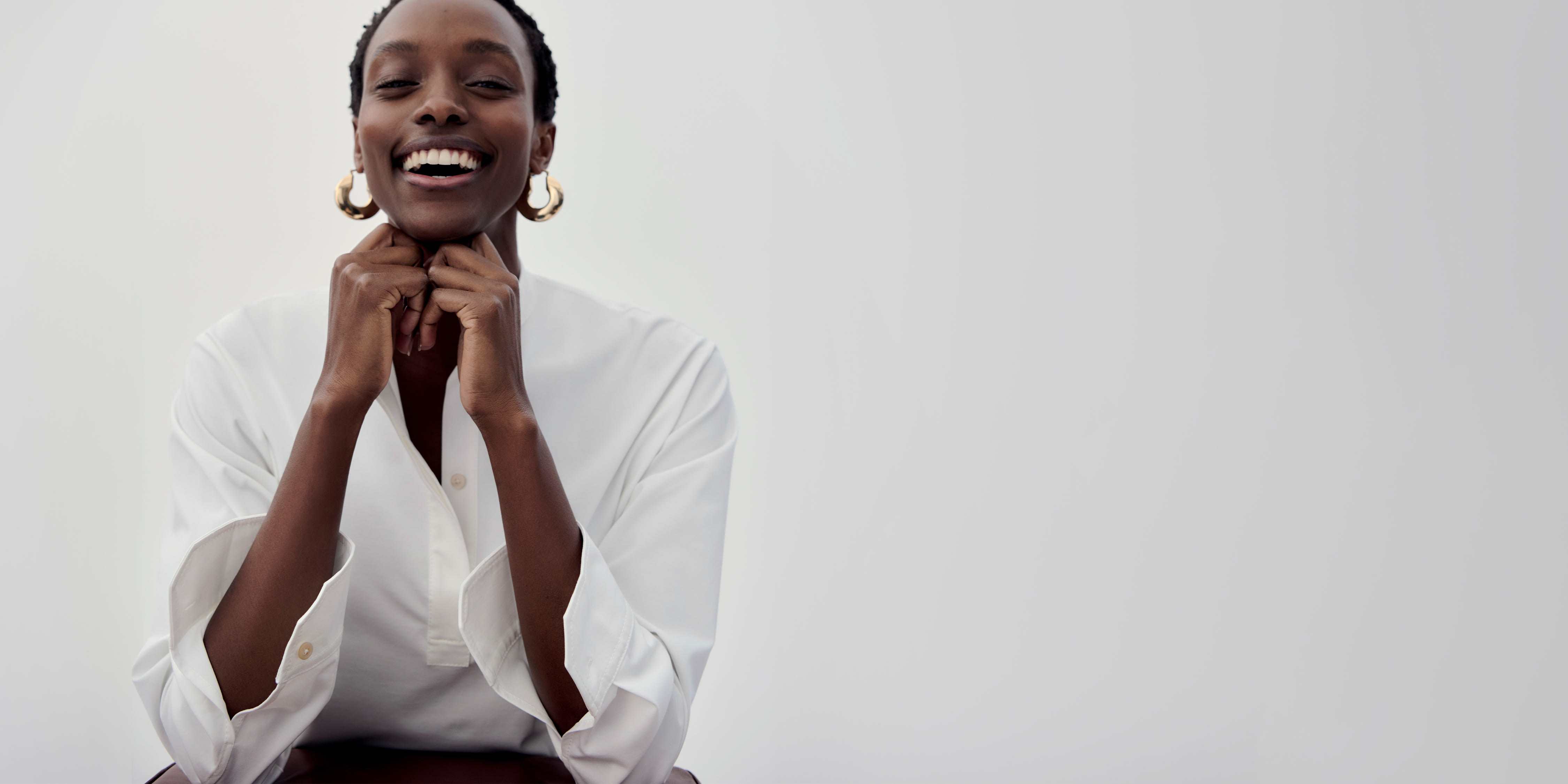 left: image of woman wearing knit poplin white button-down top and check knit suit pants, leaning on yellow pillow on wooden bench; right: woman in front of light grey background, wearing white knit poplin tunic top, resting chin on her hands and smiling