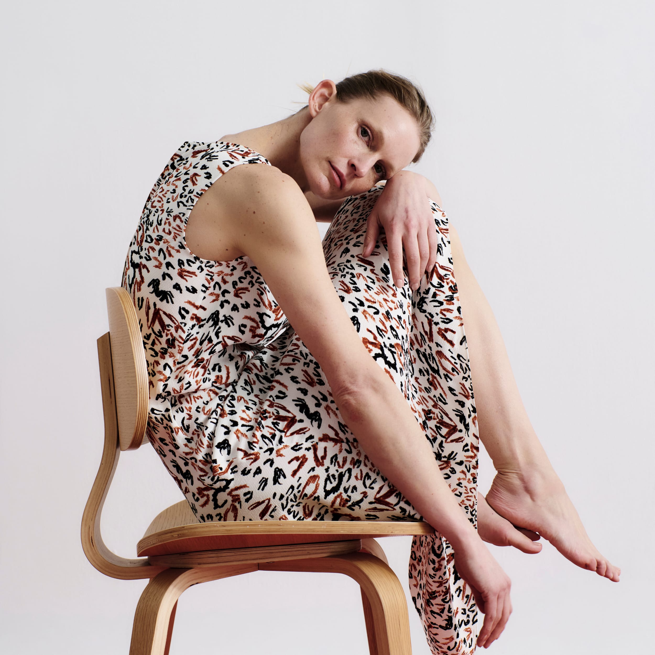 woman wearing sleeveless Priya dress in leopard sketch print, crouching with her knees to her face in a chair