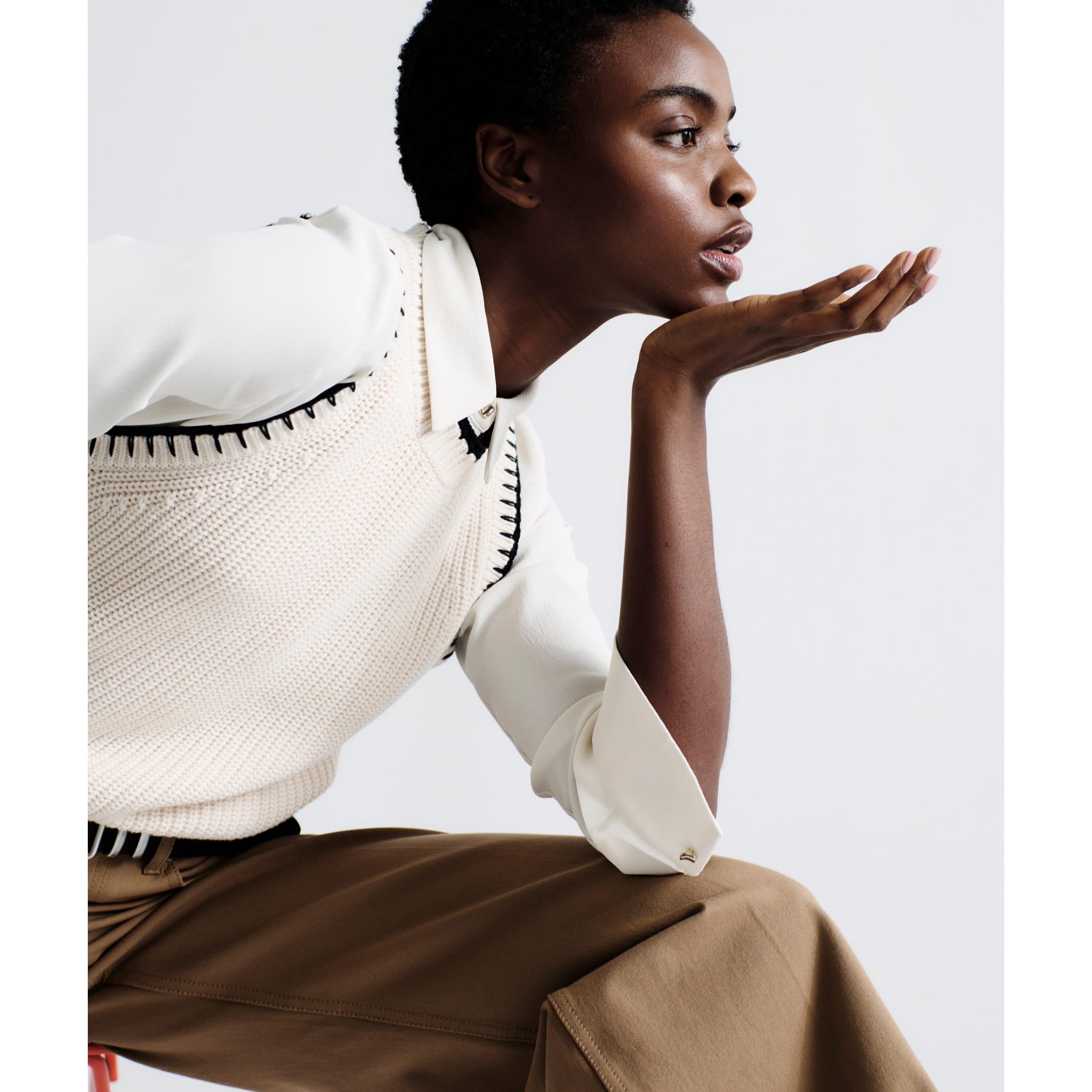 woman resting elbow on one leg and chin in hand, wearing white button-up shirt under white knit Giada sleeveless top with black contrast stitching