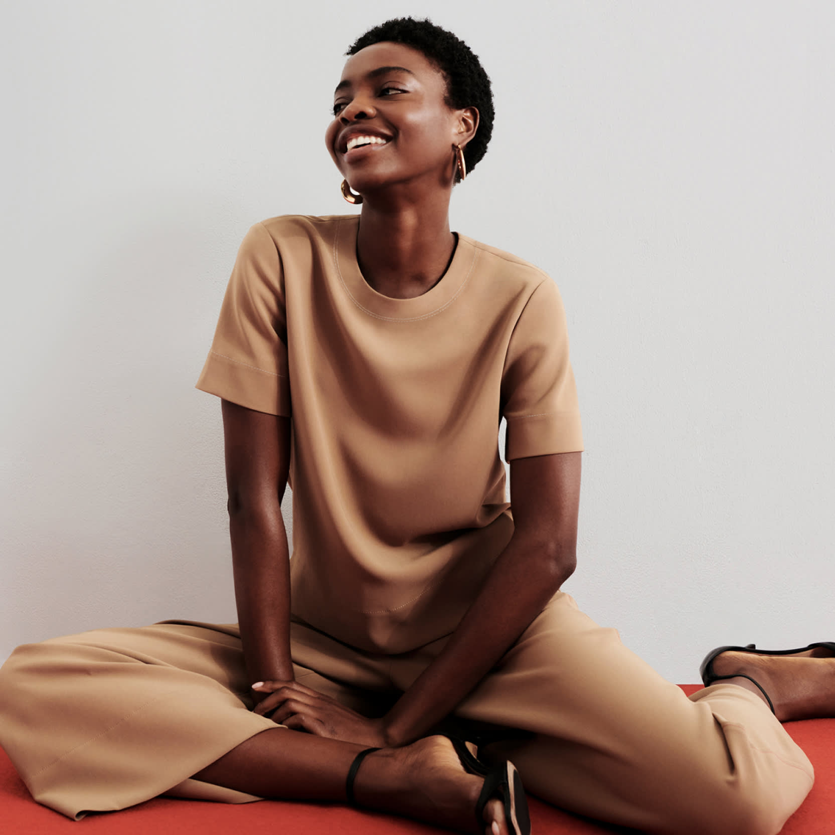 woman sitting on red floor wearing matching saddle-colored short sleeve top and culottes, smiling