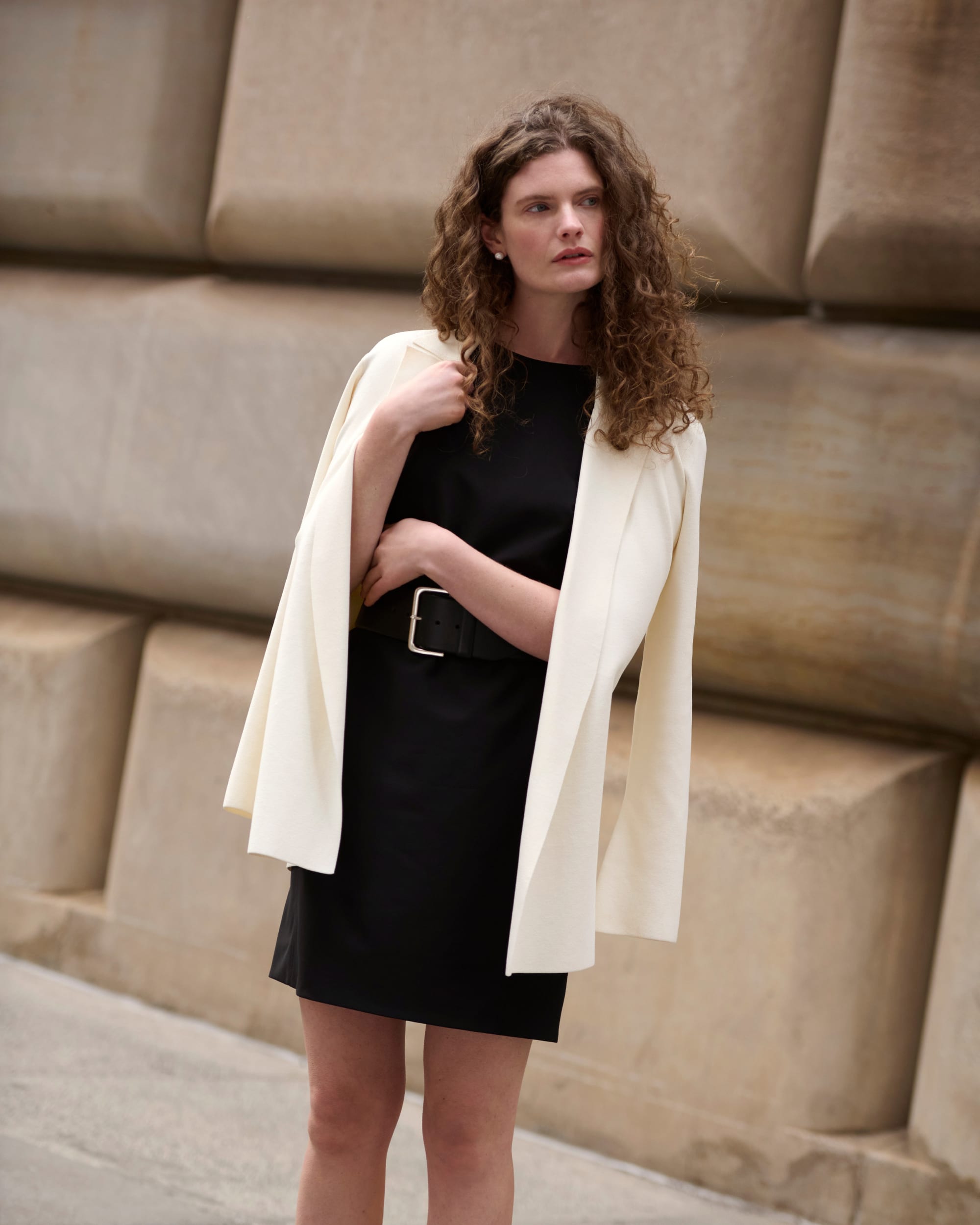 image of a woman wearing the merritt jardigan in ivory over the maaza dress in black standing on the sidewalk