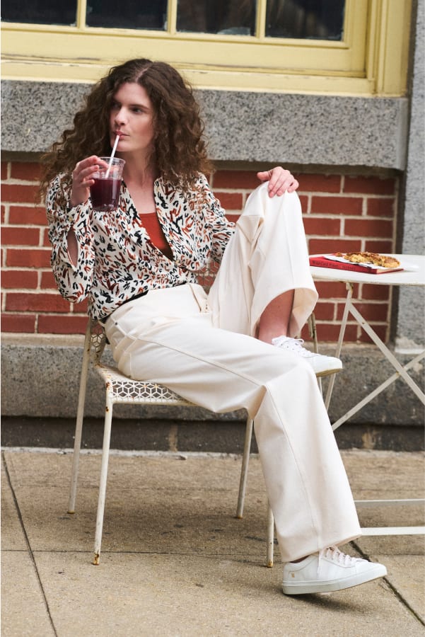 woman sitting with one leg up on her chair, sipping a drink and looking to the side, wearing lagarde shirt in leopard sketch print and wide-leg white milo jeans and white sneakers