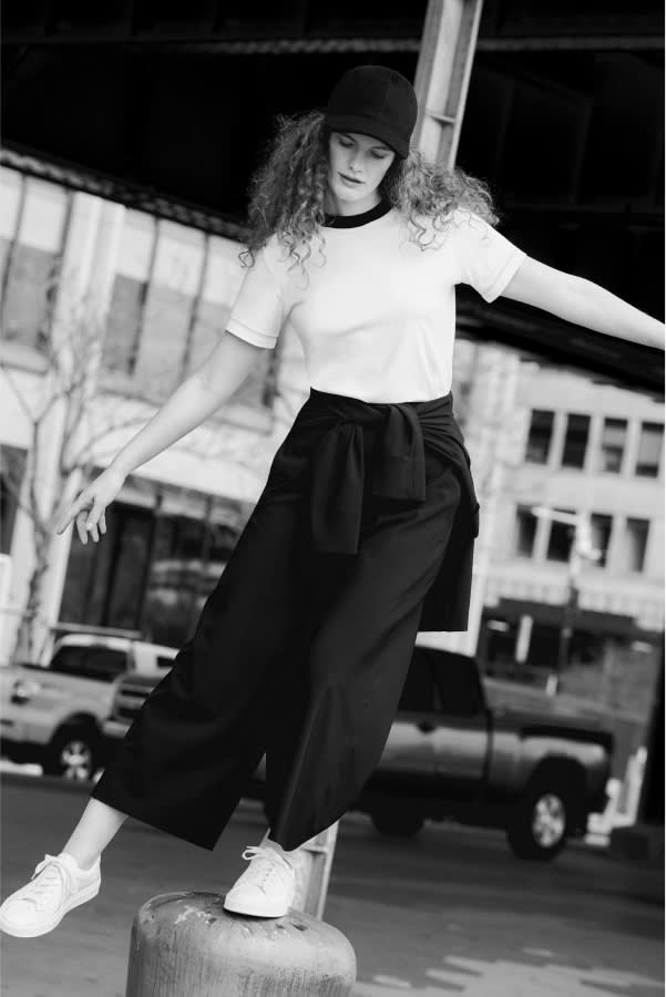 black and white image of woman wearing white T-shirt with black contrast collar, wide-leg black culottes, and black baseball cap, balancing on pedestal outside